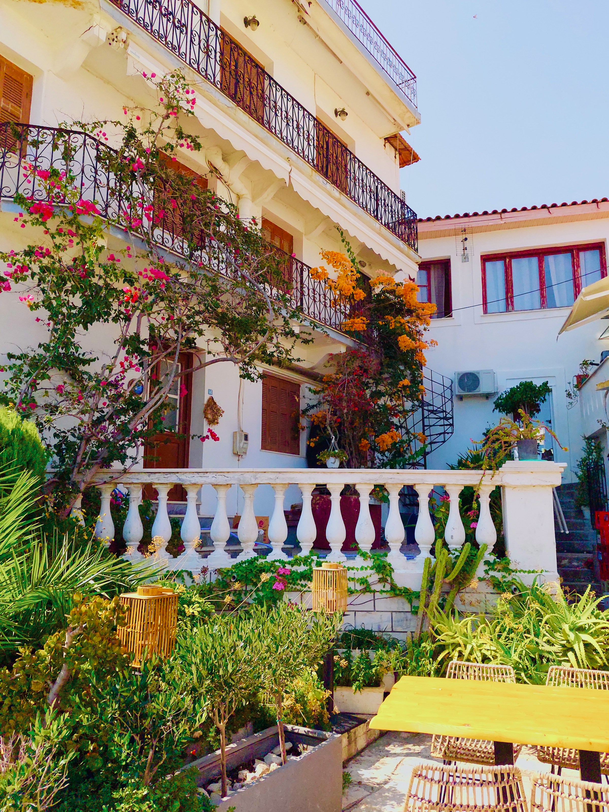 Charming Mediterranean building with flowering plants, wrought iron balconies, and lush greenery under clear blue sky.