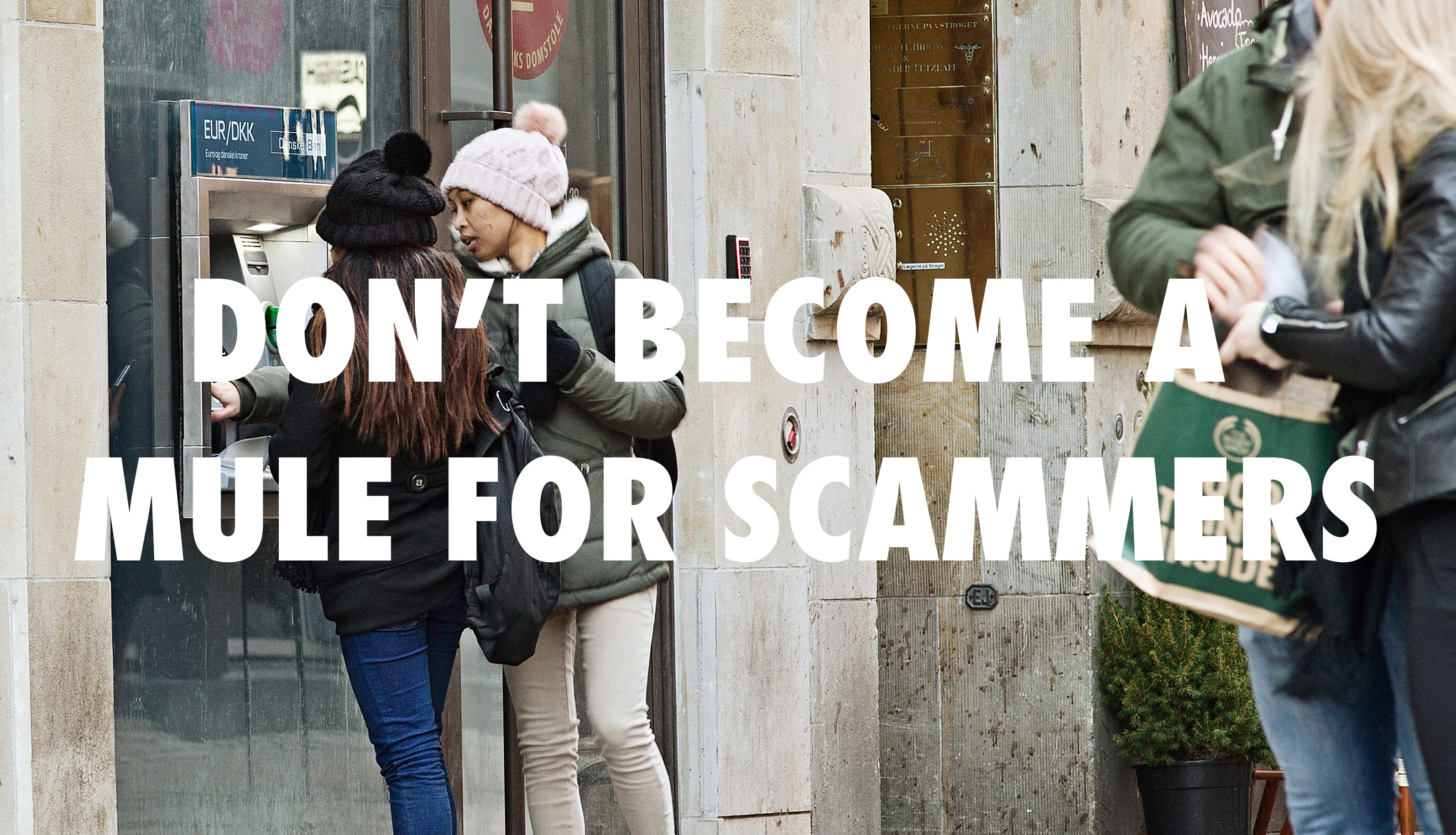 Personer ved en hæveautomat i byen med teksten "DON'T BECOME A MULE FOR SCAMMERS", advarer mod bedrageri
