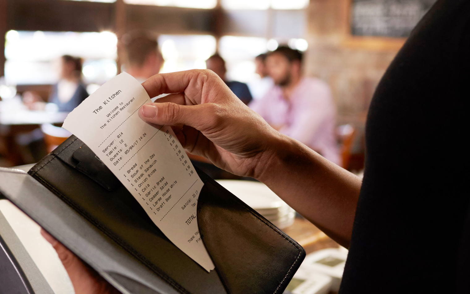 Person, der holder en restaurantregning i en læderholder, mens han sidder ved et bord på en restaurant. Flere personer kan ses i baggrunden, samt et sløret restaurantinteriør.