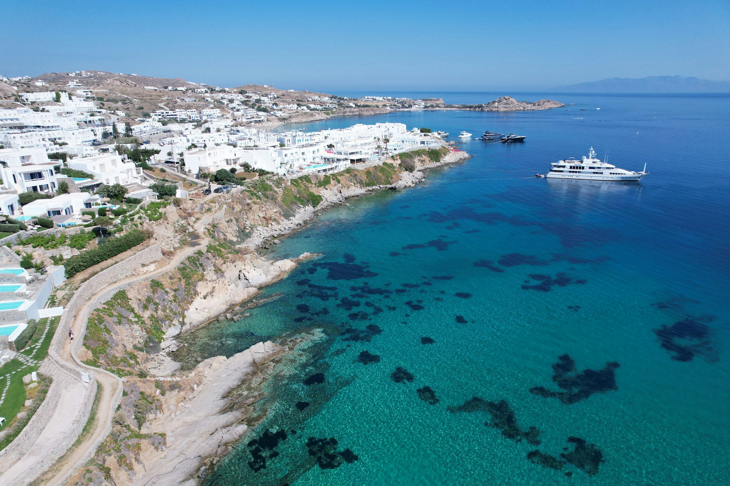 Luftfoto af kysten af Mykonos, Grækenland, med turkisblåt hav, hvide bygninger og både ved havnen.