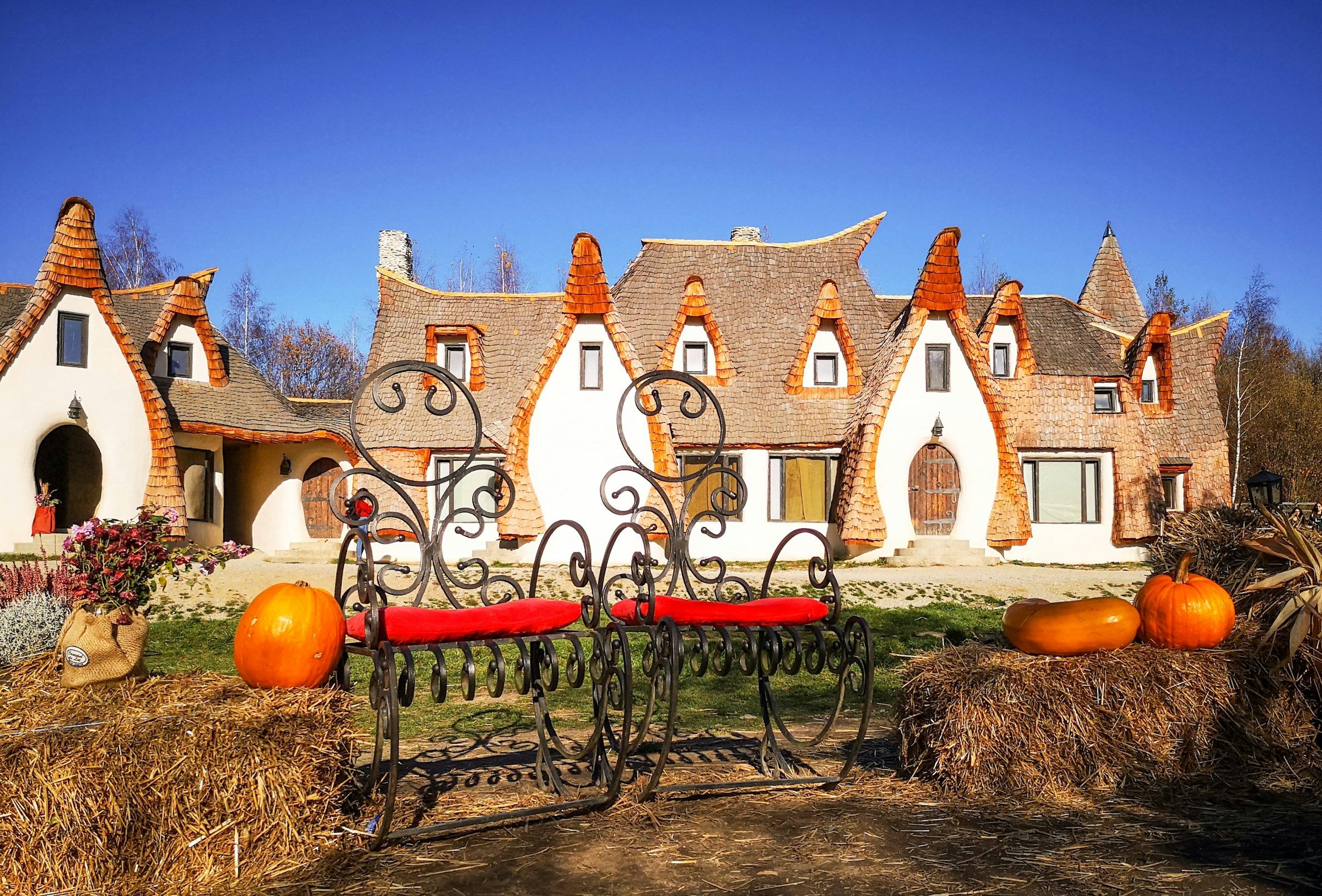 Fabelagtig arkitektur med spirede tage af et usædvanligt hus, smedejernsbænke og efterårsgræskar foran bygningen under en klar blå himmel.