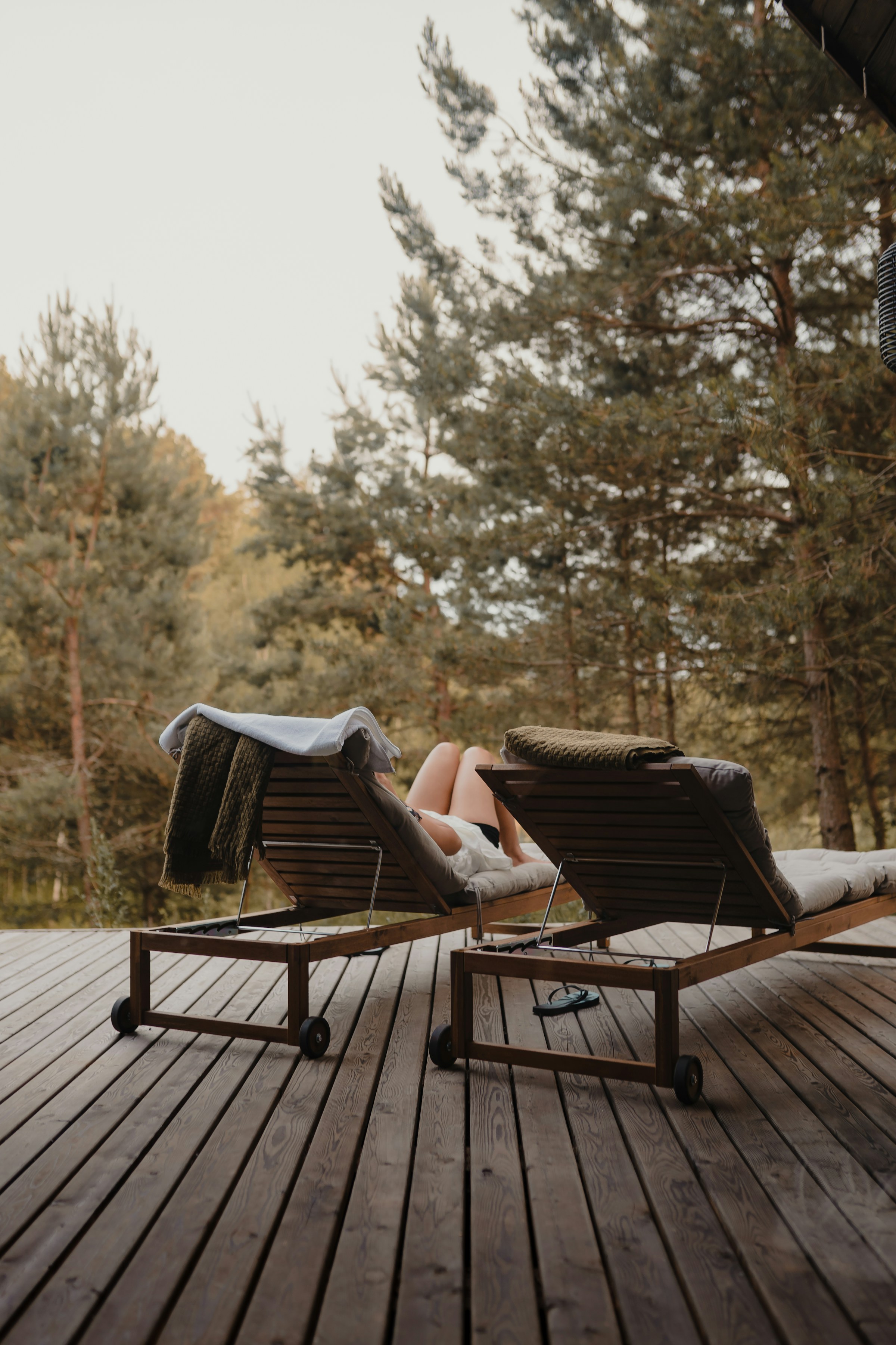 Two sun loungers on a wooden terrace surrounded by pine trees in a quiet forest environment.