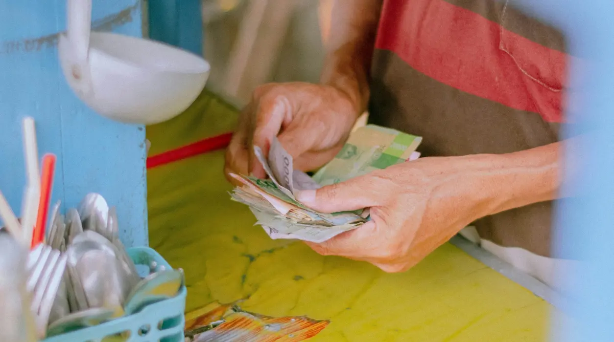 Person räknar en bunt sedlar vid en disk, med skedar och köksredskap i en behållare bredvid.