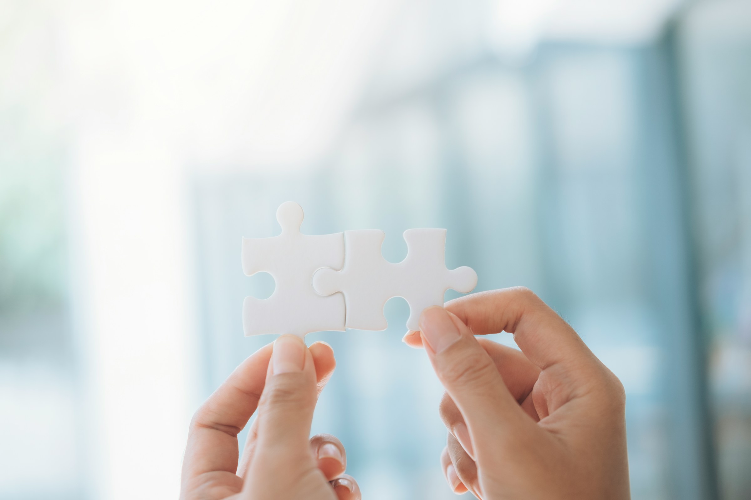 Hands holding two white puzzle pieces against a blurred background, symbolizing problem-solving and teamwork.