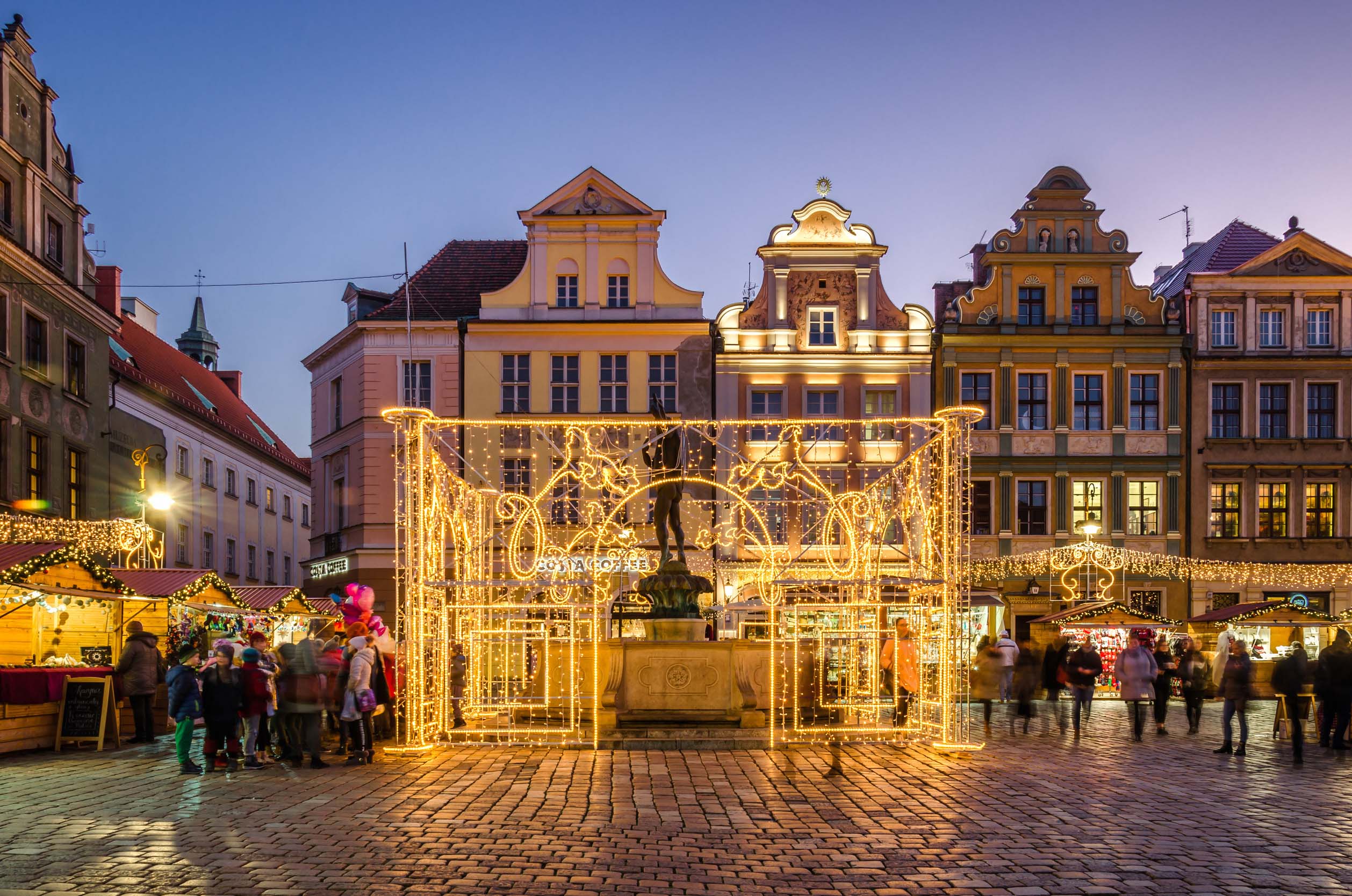 Poznań Christmas Market with light displays, market stalls, and historic buildings illuminated at dusk.