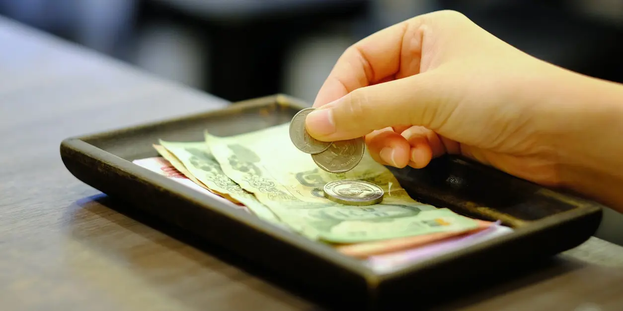 Hand lägger mynt på en bricka med sedlar och mynt, troligen som betalning eller dricks på en restaurang.