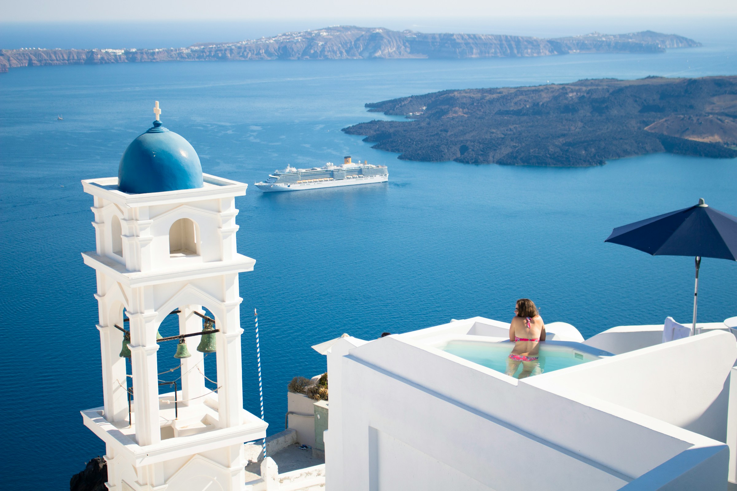 Kvinde slapper af i en infinity-pool med udsigt over havet med et krydstogtskib og en blåkupplet kirke i Santorini, Grækenland