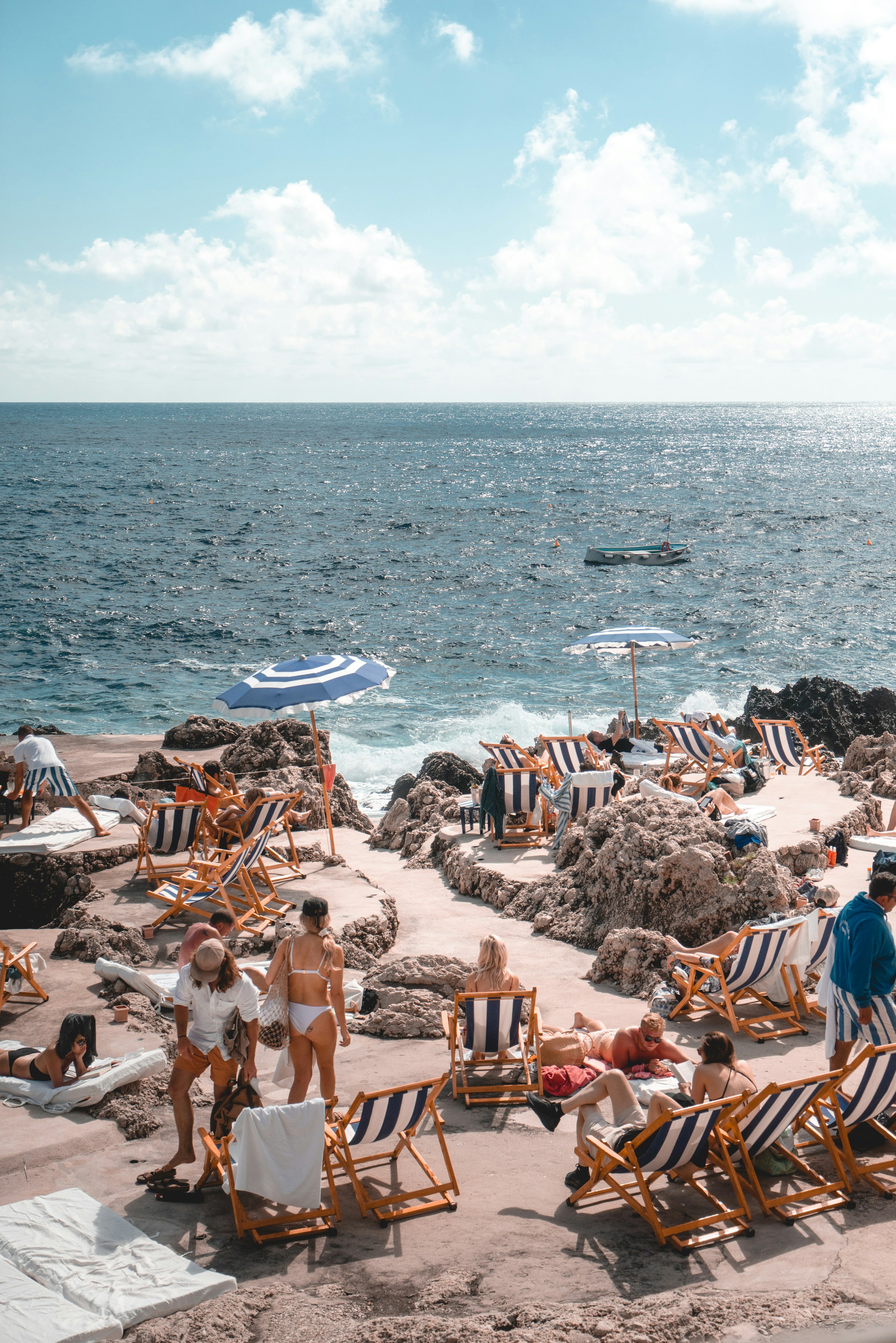 Solstole og parasoller på den klippefyldte kyst med turkis hav og badende, der nyder strandferie ved Middelhavet. Afslappende strandmiljø i Sydeuropa.