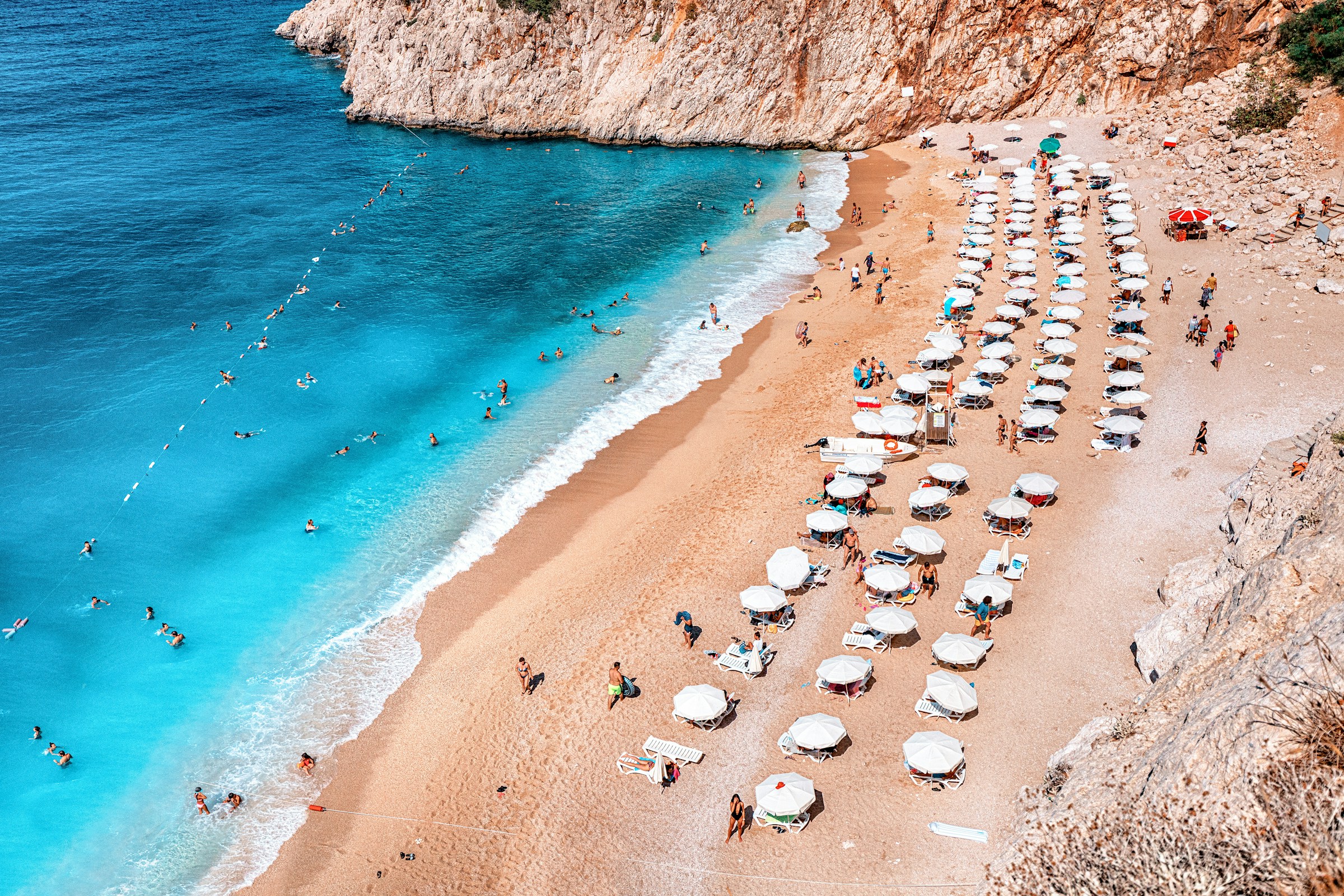 Sandy beach with rows of white umbrellas and sun loungers, blue ocean waves, and people swimming and relaxing near rocky cliffs.