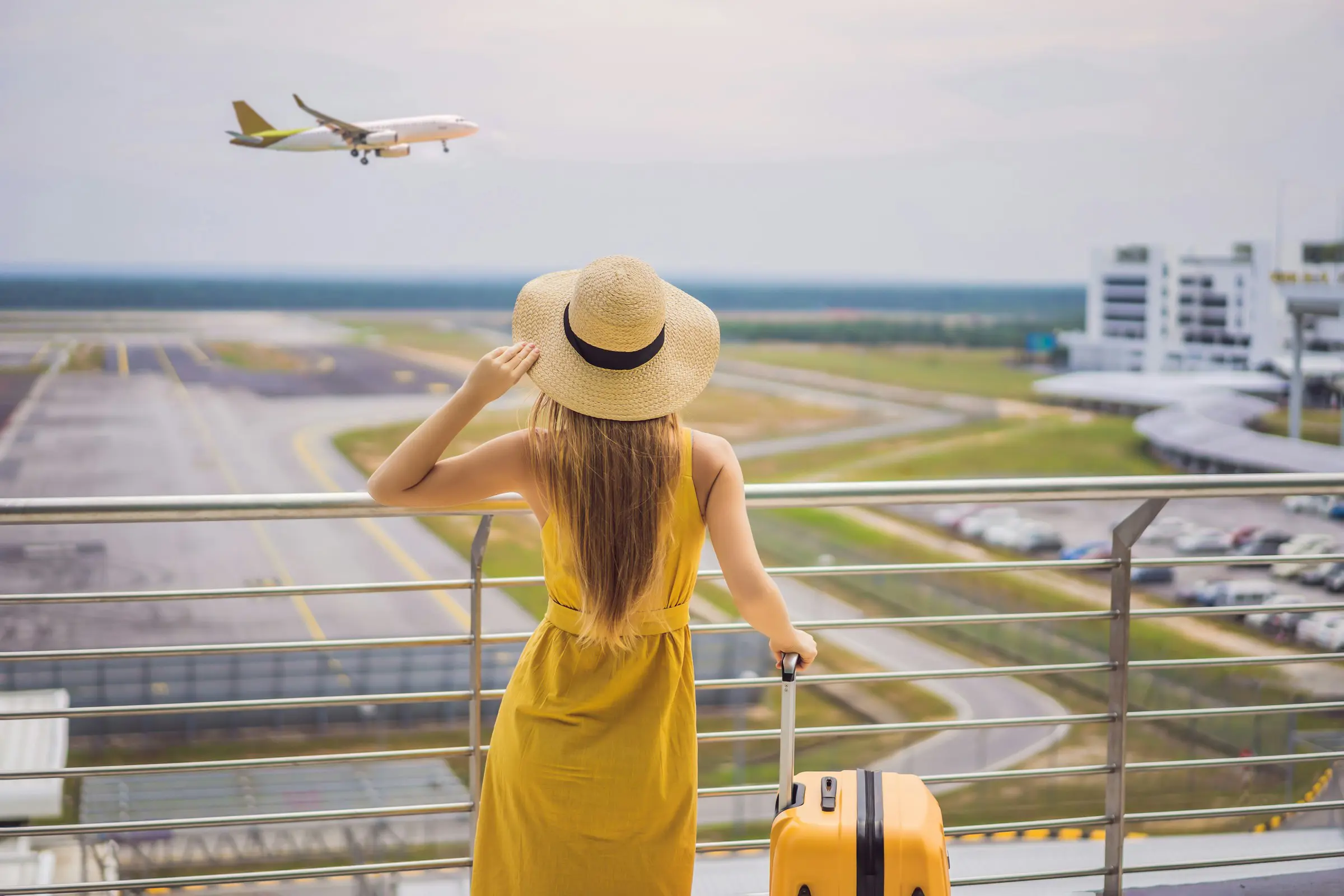 Kvinde i gul kjole og stråhat ser på fly i lufthavn, mens hun står ved rækværk med gul kuffert