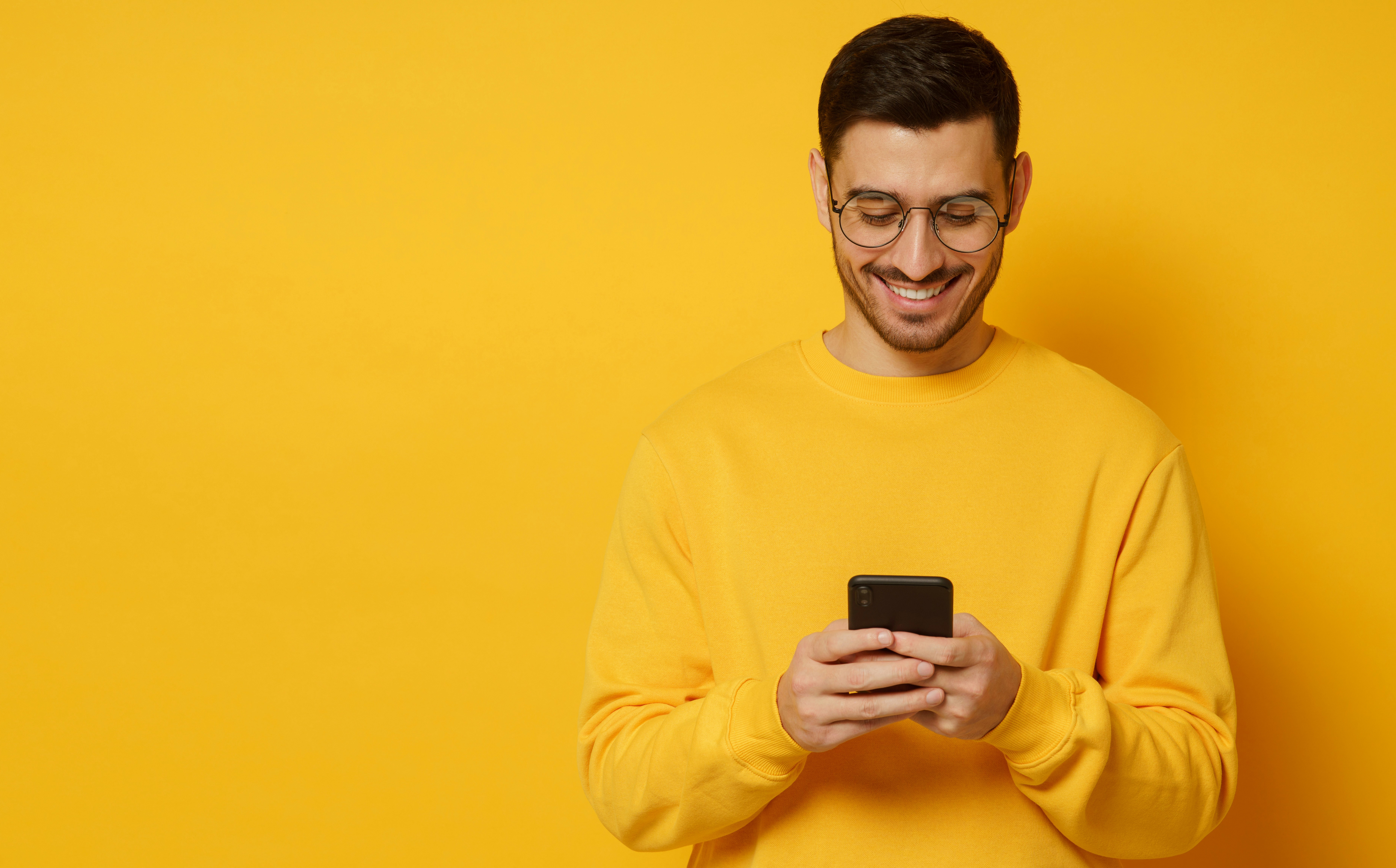 Smiling man in glasses wearing a yellow sweater using a smartphone against a yellow background.
