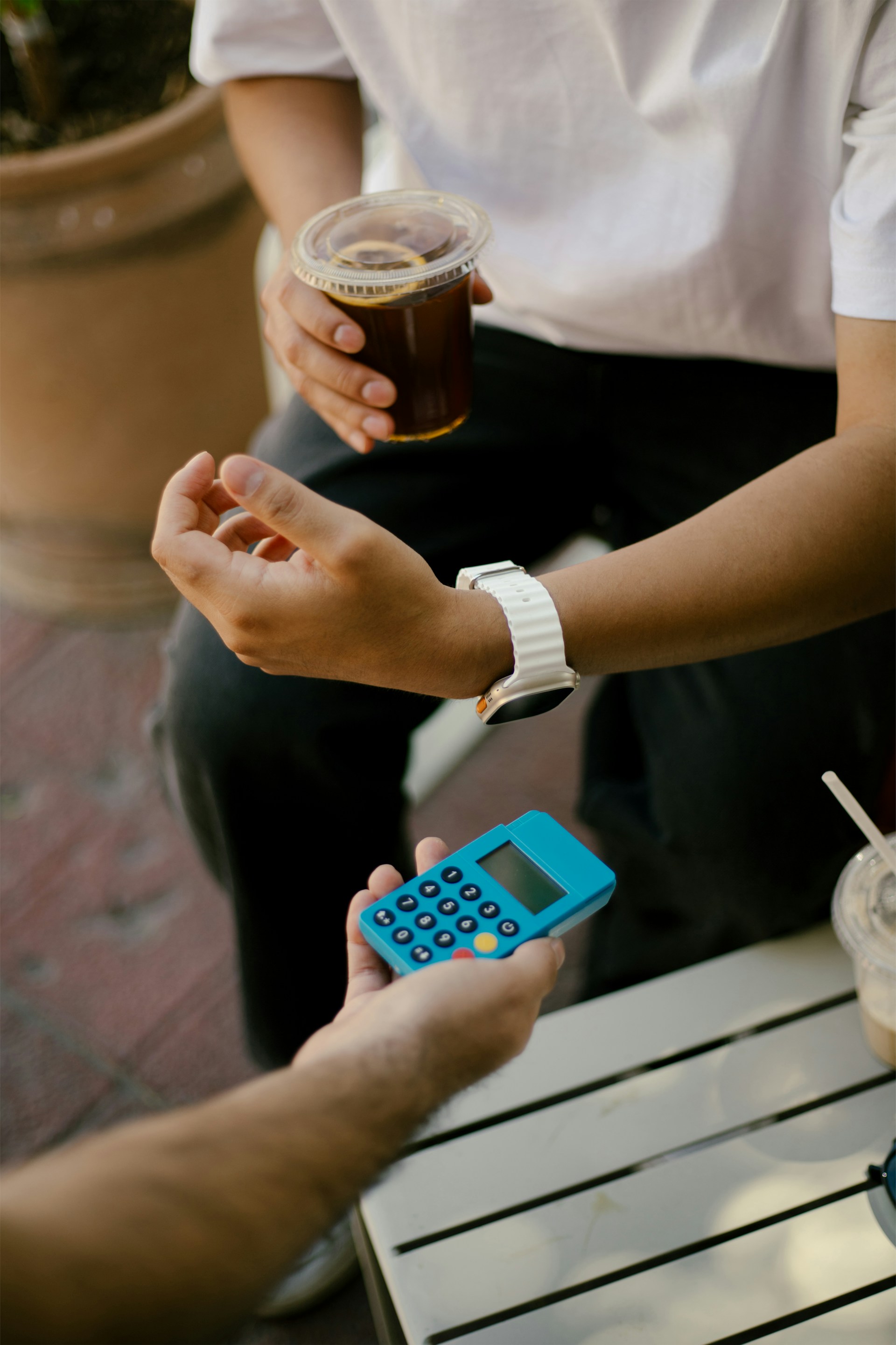 Person paying with a blue card reader while holding a cold drink in a plastic cup, sitting at an outdoor table.
