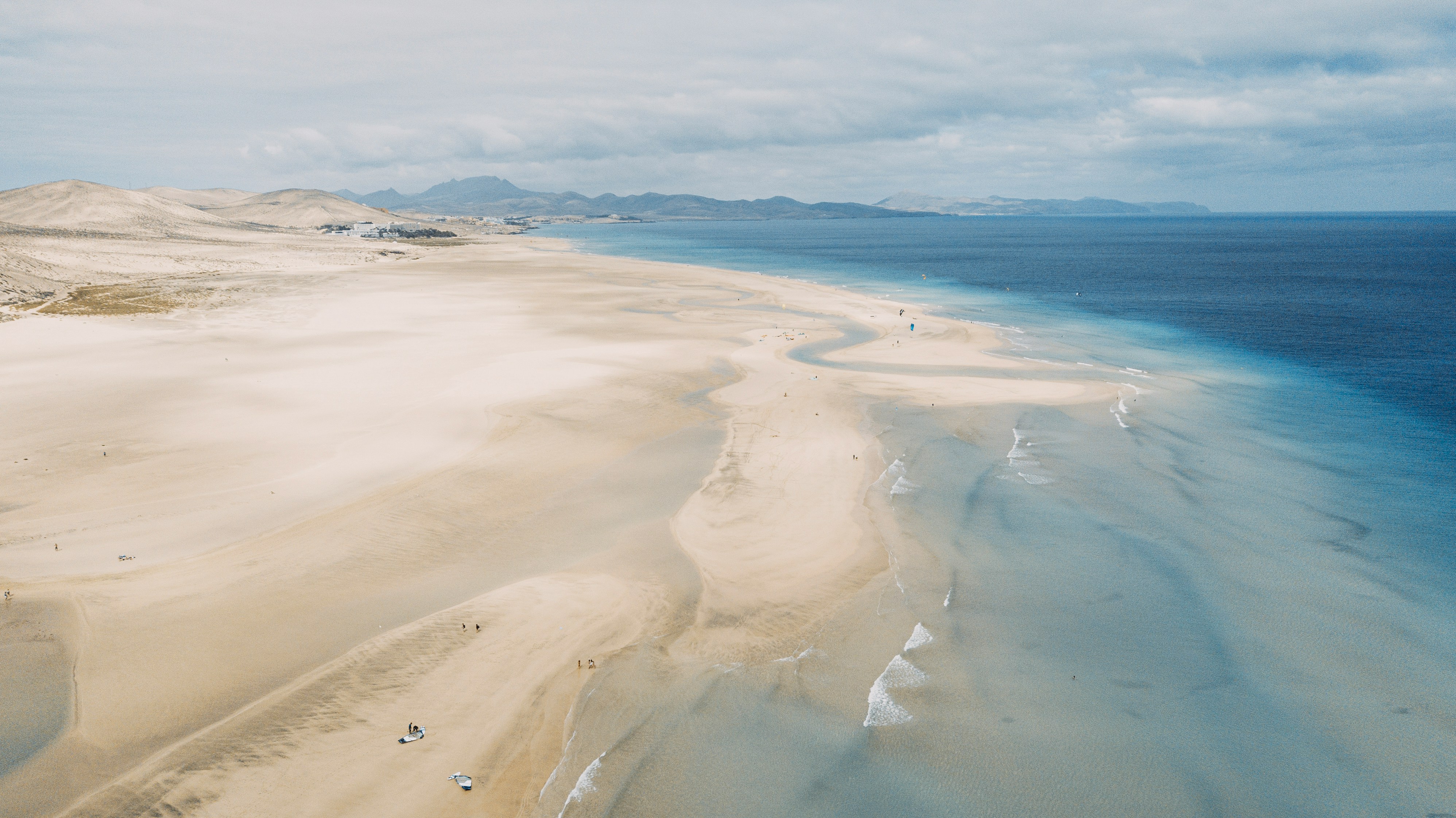 Luftfoto af Jandía-stranden på Fuerteventura, med gyldent sand og turkisblå hav. Forrevne bjerge ses i baggrunden under en overskyet himmel. Perfekt til strandferie