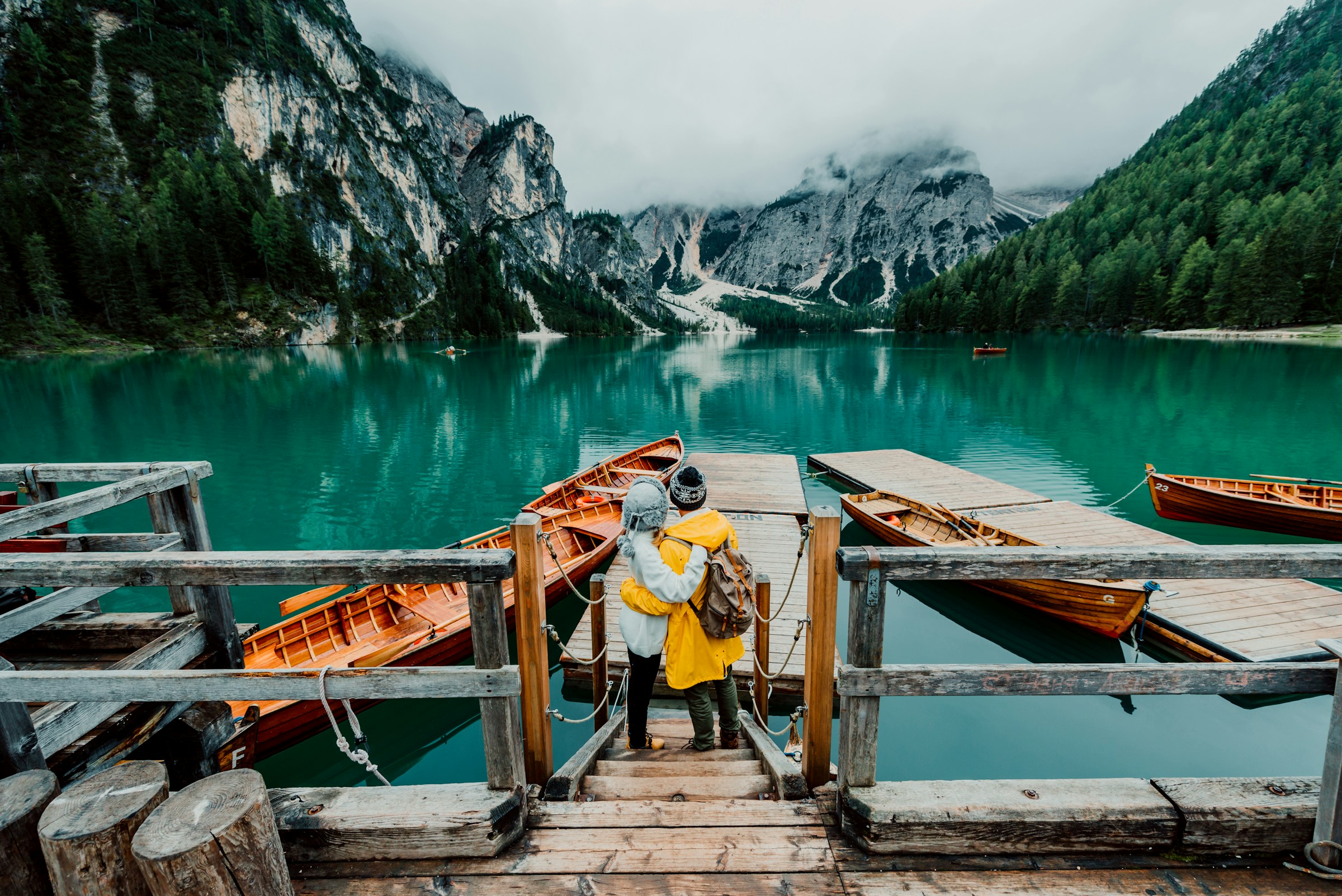 Couple embracing on a wooden dock overlooking turquoise lake and mountains in a foggy landscape.
