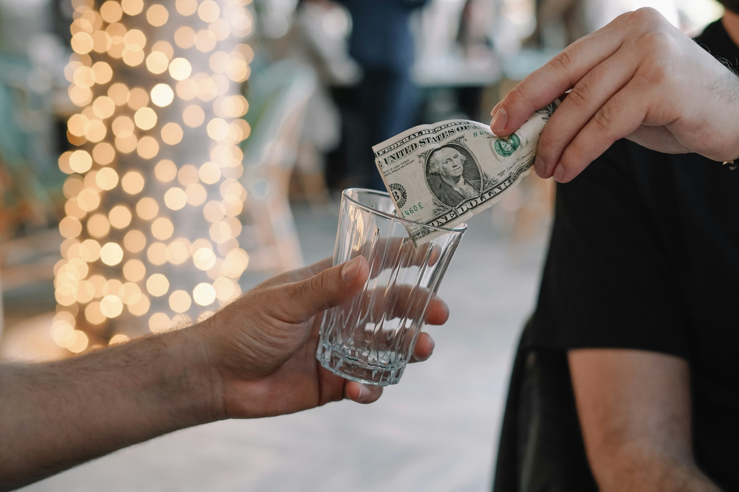 Person placing a dollar bill into a glass against a backdrop of blurred decorative lights, symbolizing tipping or donation in a festive setting.