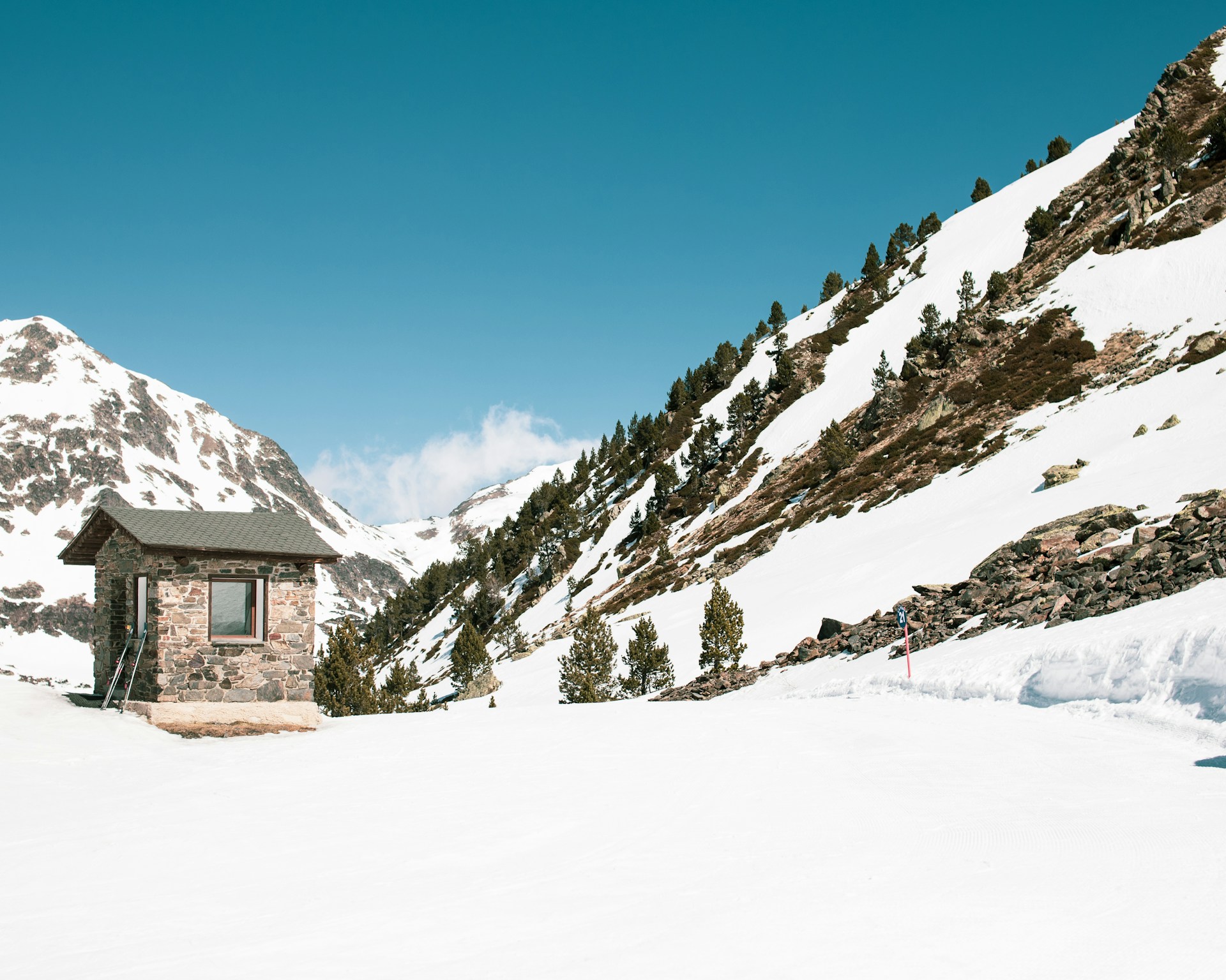 Stone cabin in snowy mountain landscape with evergreen trees under a clear blue sky.