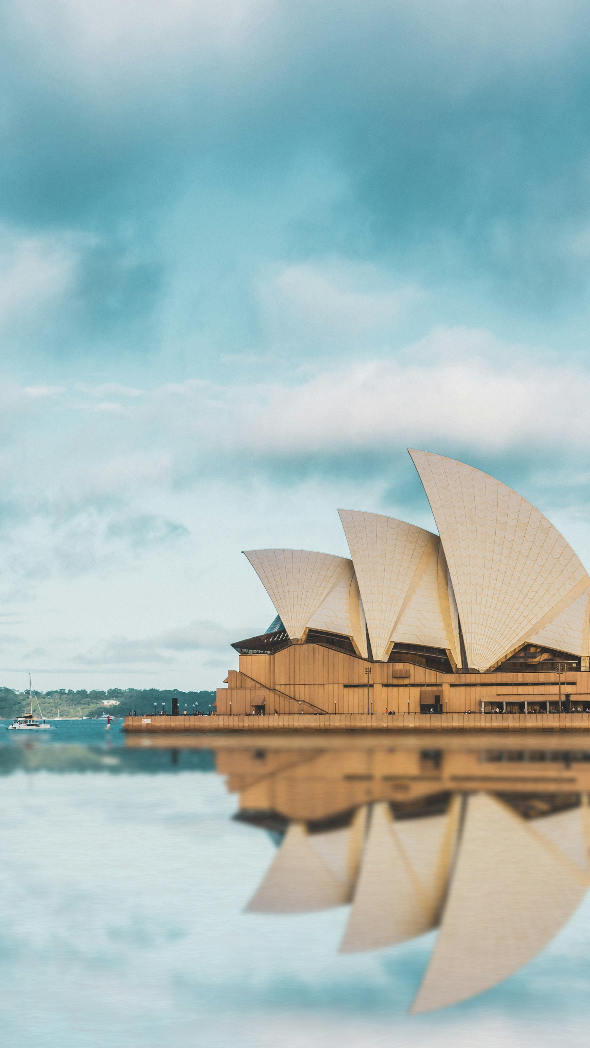 Sydney Opera House with its iconic sails reflected in calm water under a partly cloudy sky.