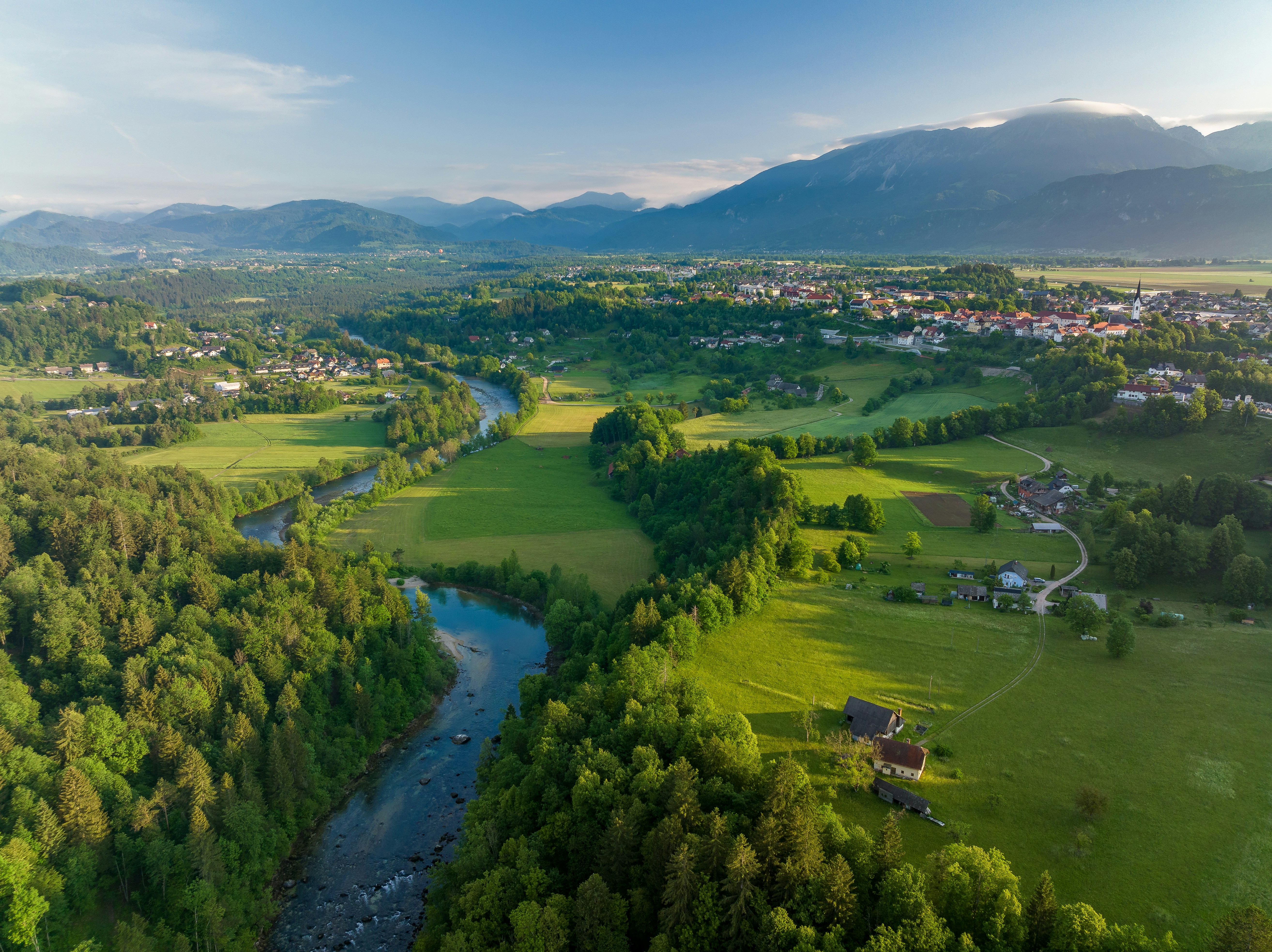 Luftfoto af frodigt landskab med grønne marker, flod og by, omgivet af bjerge under en klar blå himmel i dagtimerne