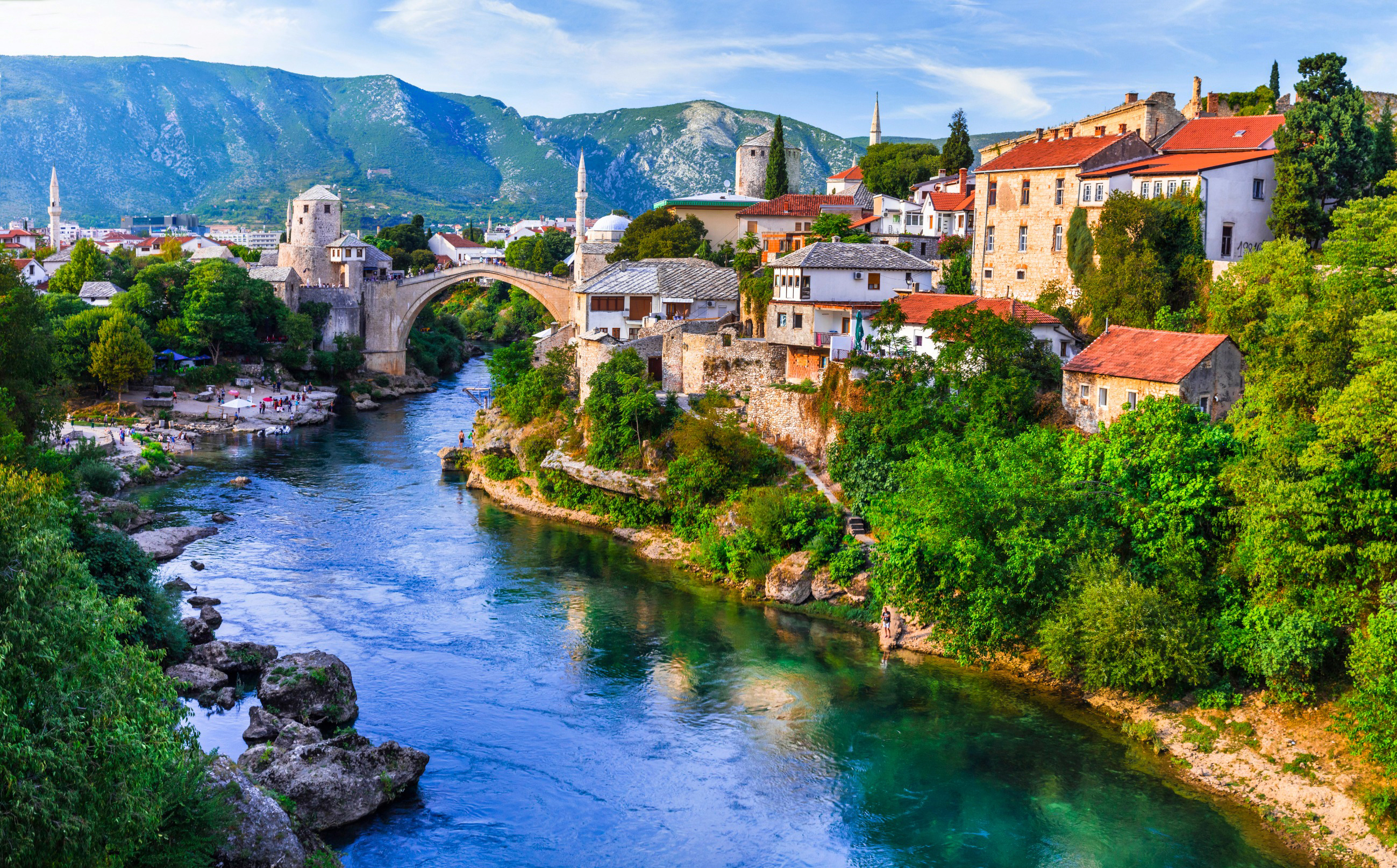 Panoramisk udsigt over Mostar med den ikoniske gamle bro, Neretva-floden og historiske bygninger, omgivet af frodig natur og bjerge i baggrunden