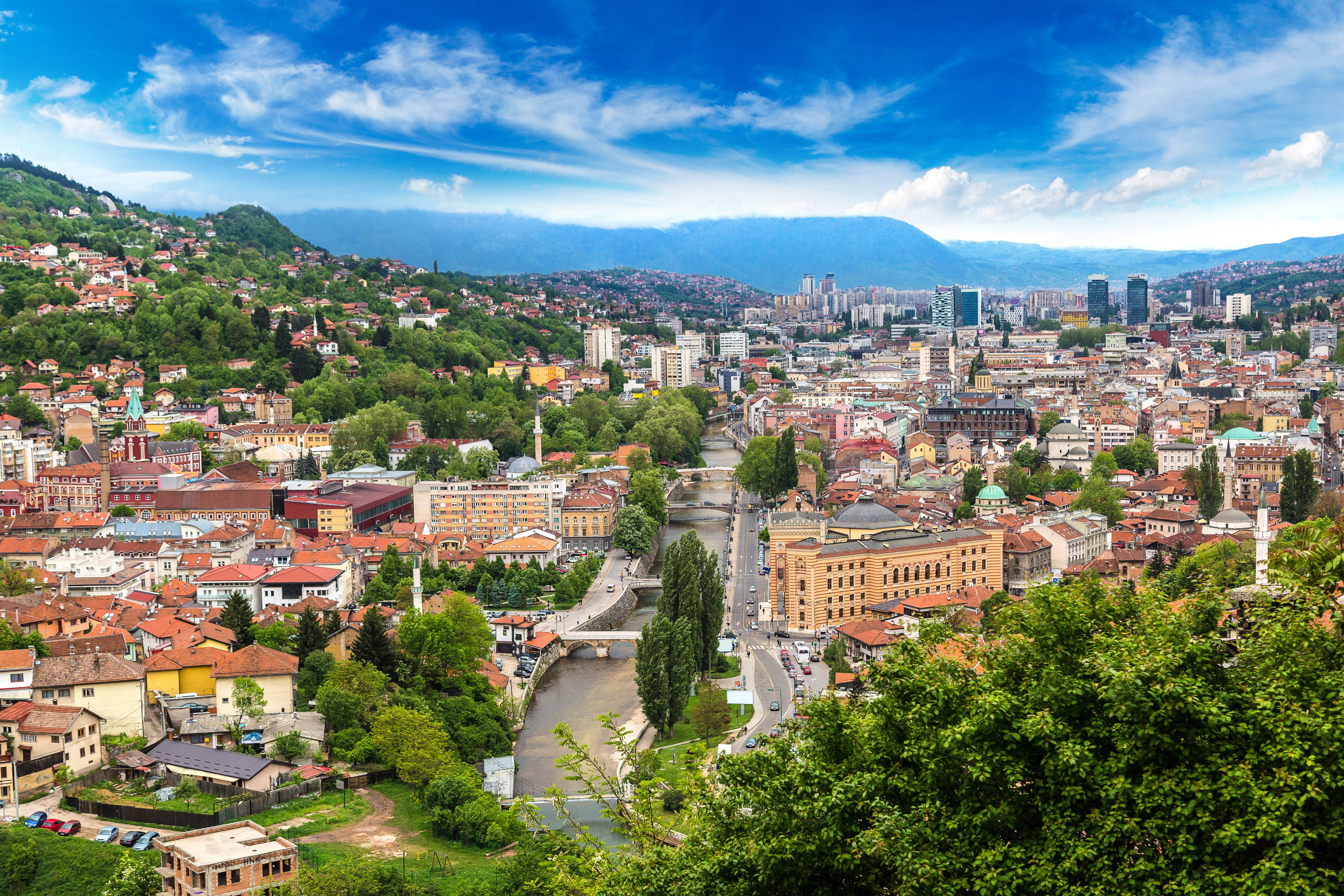 Panoramaudsigt over Sarajevo by med floden Miljacka, omkranset af grønne bakker og moderne bygninger