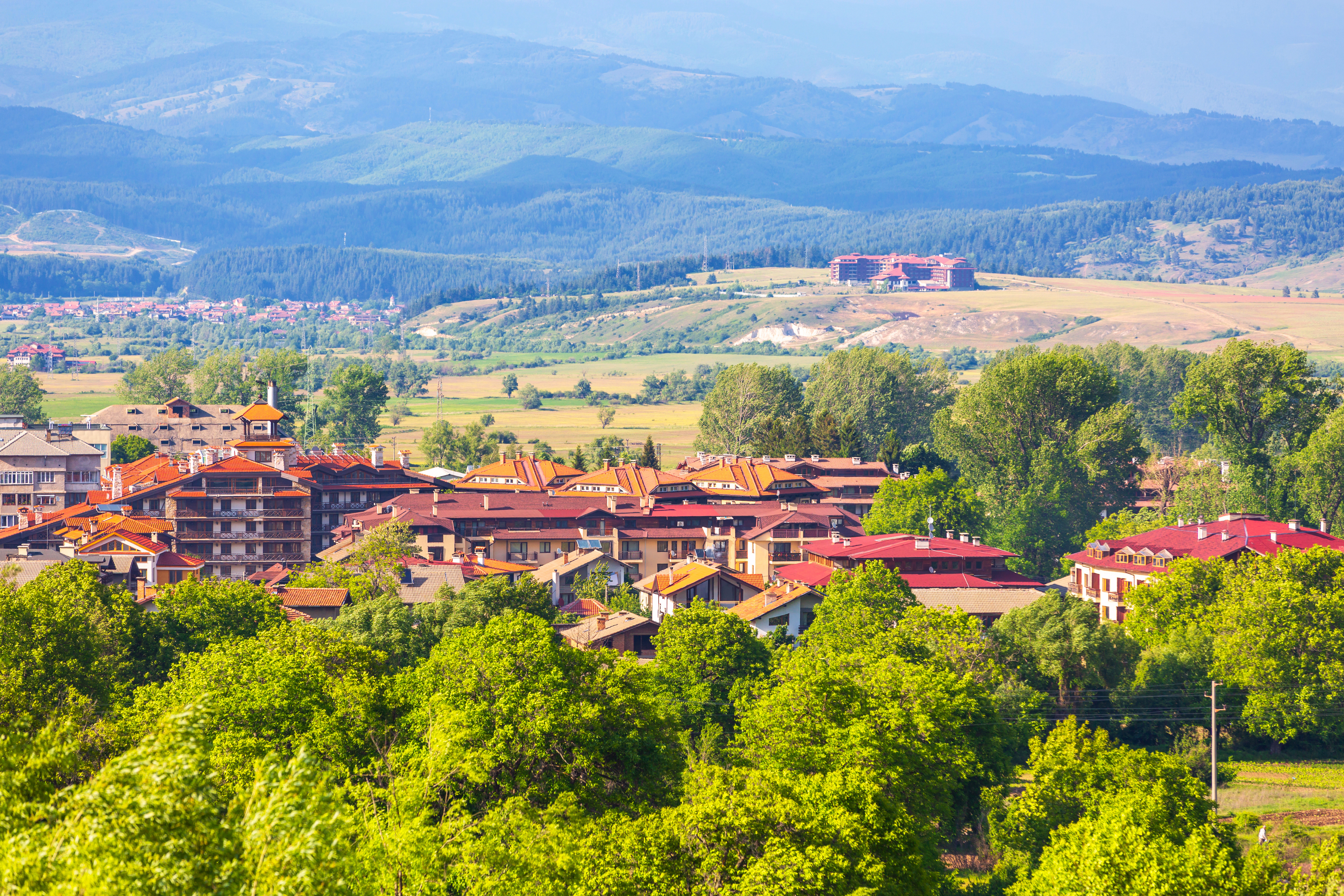 Landsby omgivet af grønne træer og bakker i forgrunden, med en bjergkæde i baggrunden på en solskinsdag