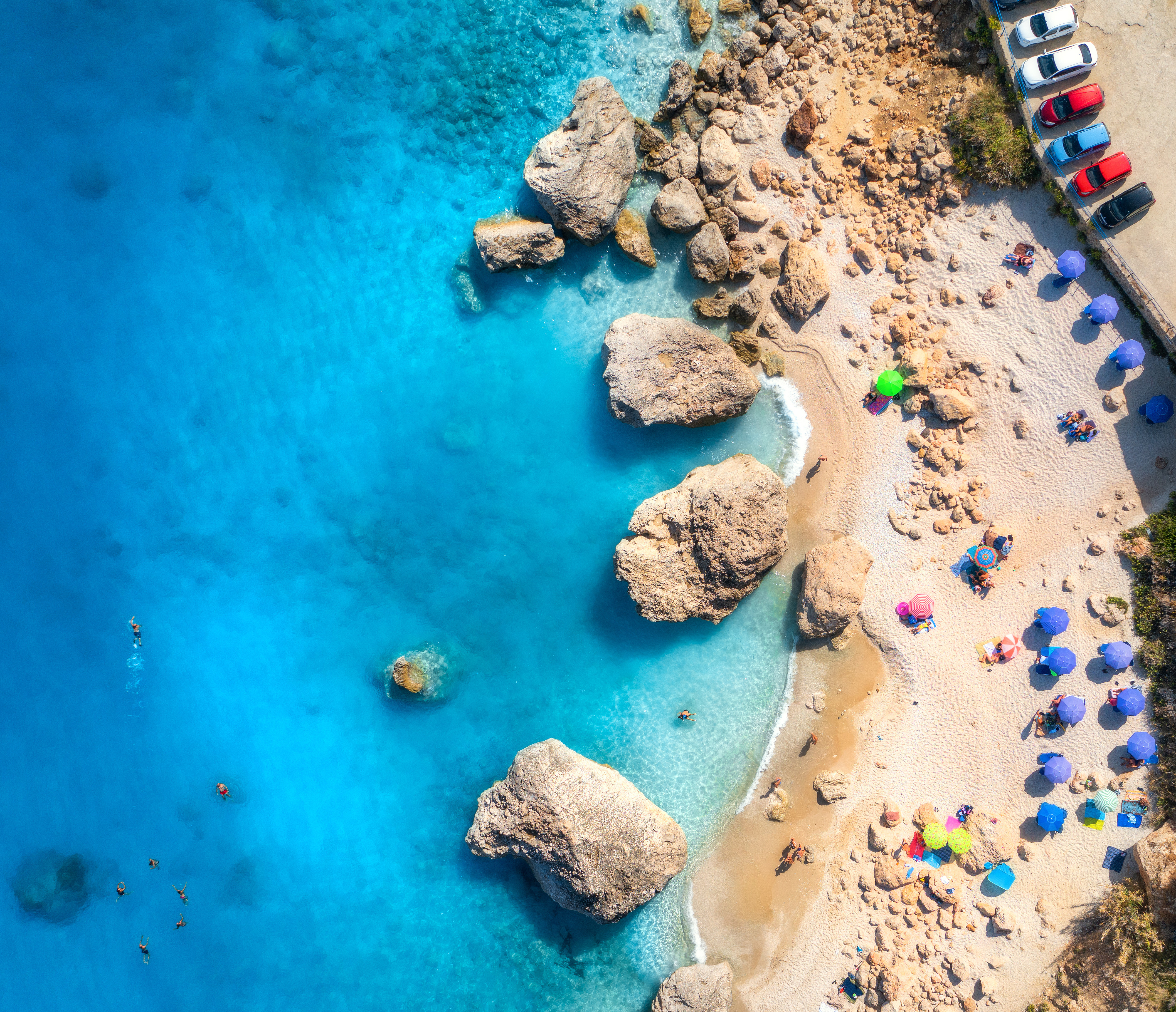 Travel to Ayia Napa - view of a picturesque beach with turquoise waters, large rocks, colorful umbrellas, and parked cars along the shore.