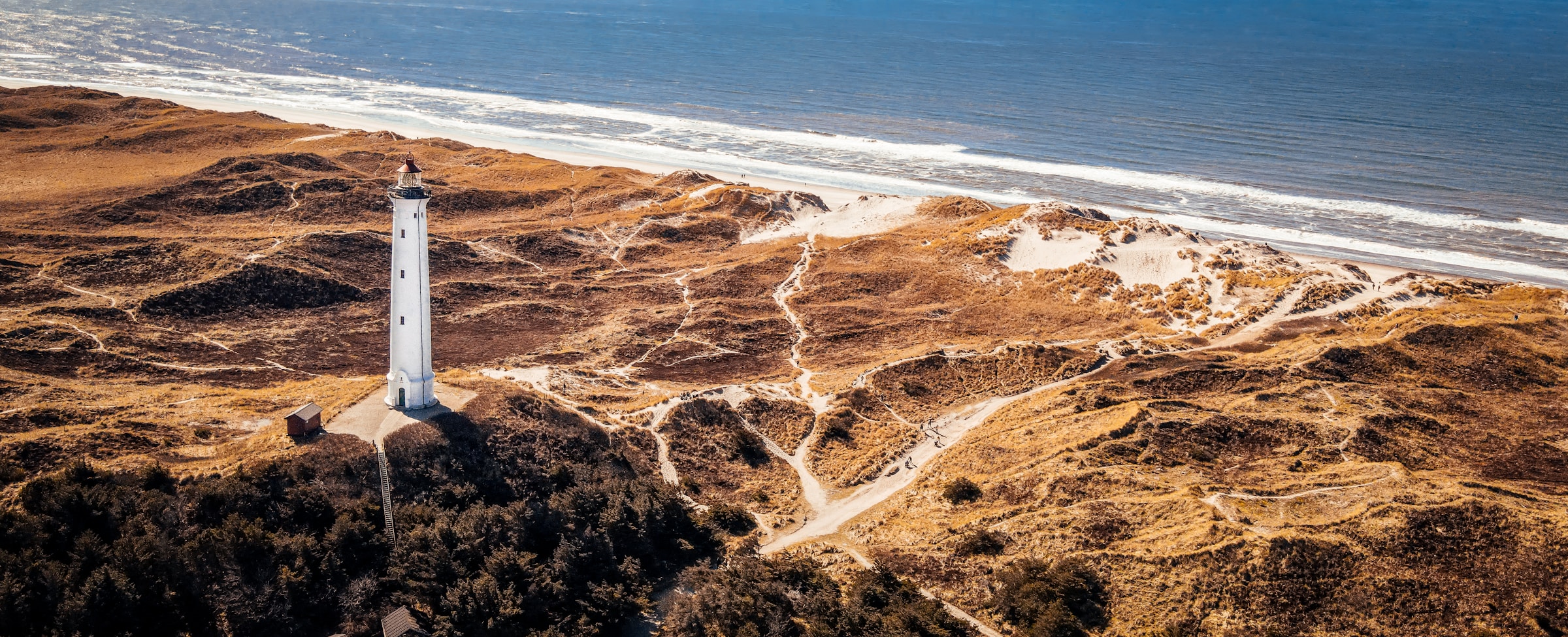 Aerial view of a coastal landscape featuring a white lighthouse surrounded by rugged dunes and vegetation, with the ocean in Denmark.