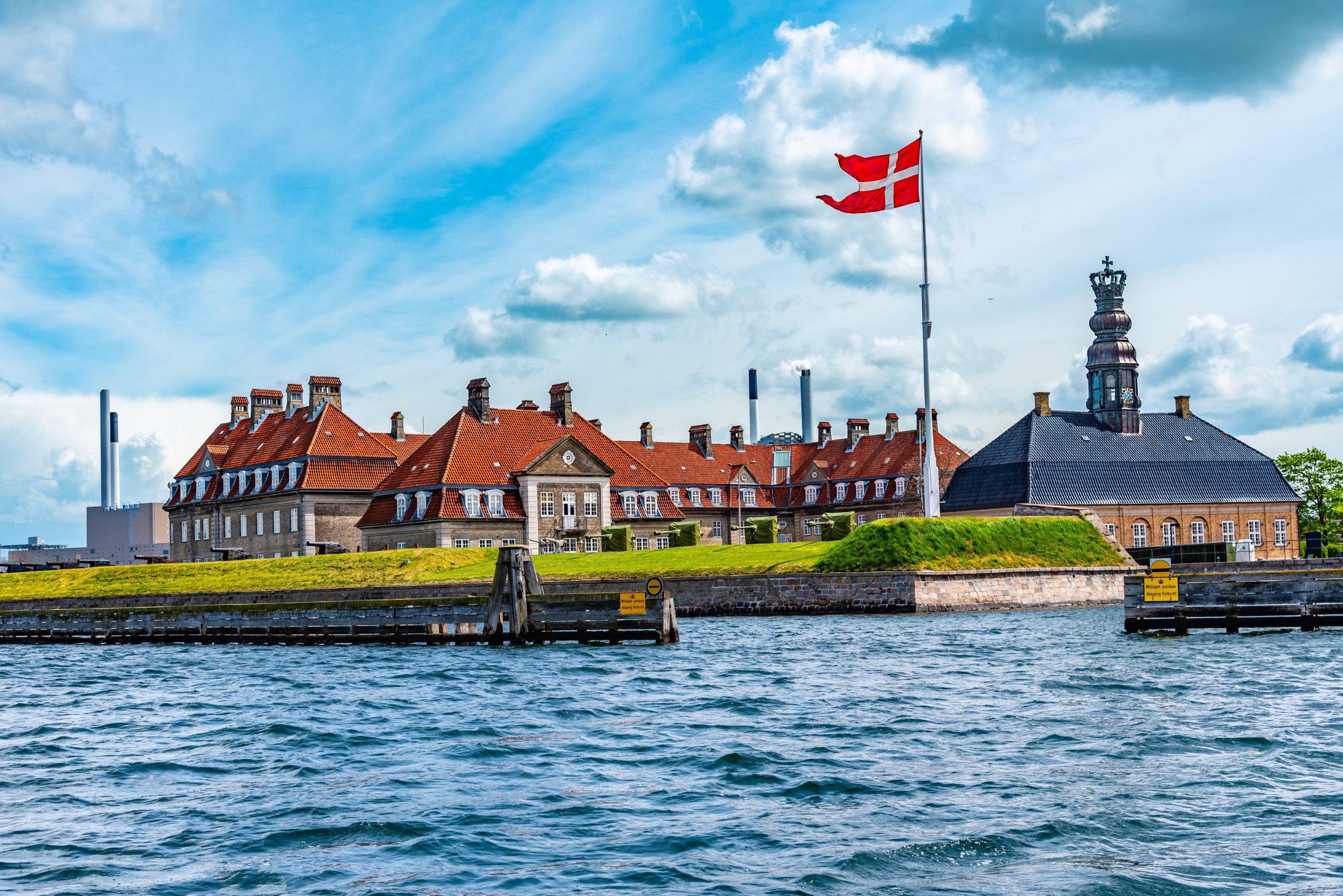 Travel to Denmark - Historic building in Copenhagen harbor with red-tiled roofs, Danish flag, and blue sky.