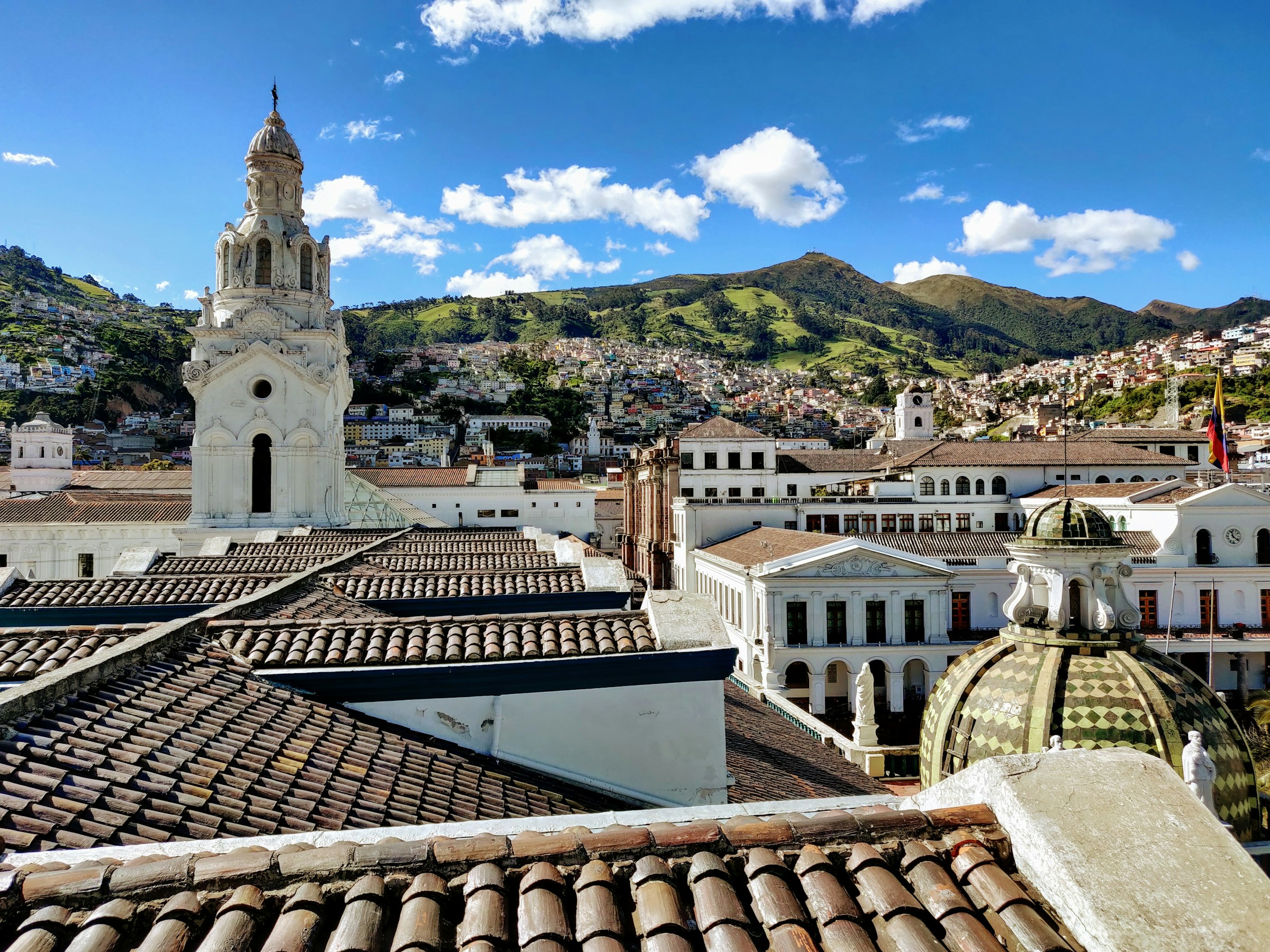 Historic Quito skyline with colonial architecture, Ecuadorian flag, and rolling green hills under a clear blue sky.