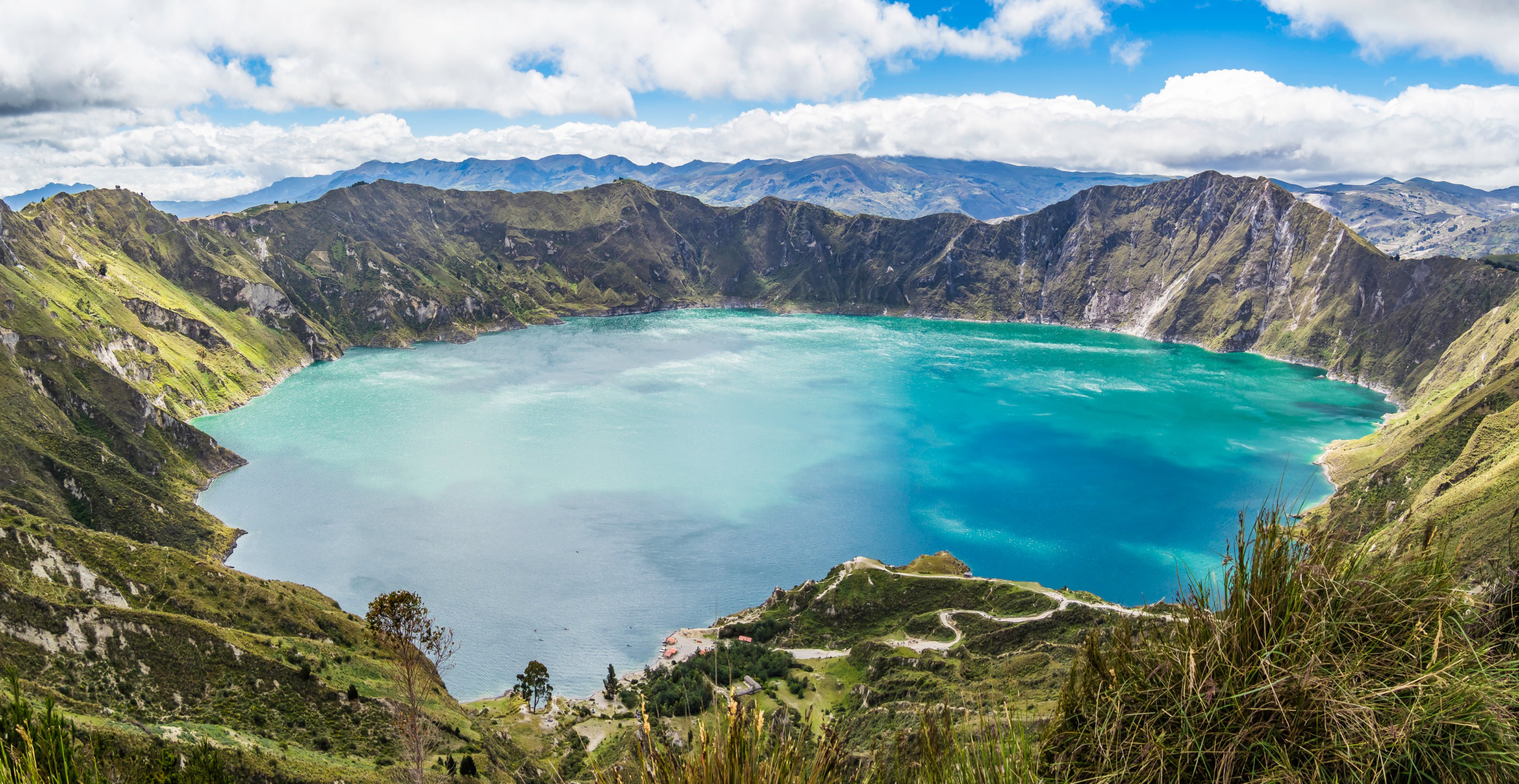 Travel Ecuador - View of Quilotoa Lake, surrounded by lush greenery and rugged cliffs under a partly cloudy sky.