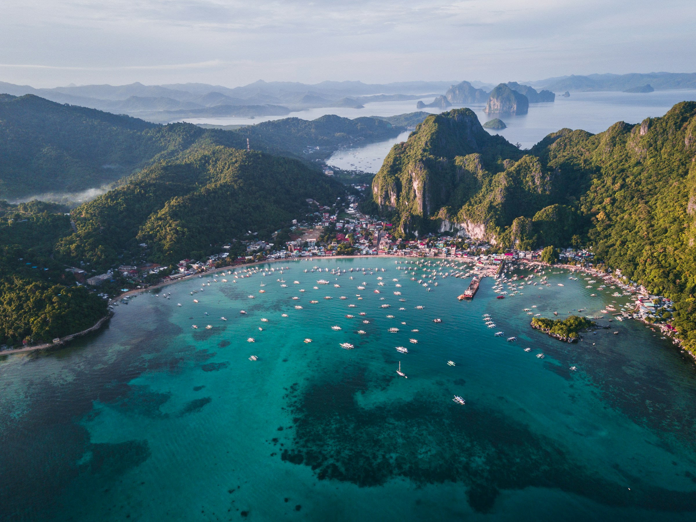 Aerial view of a coastal town in El Nido, Palawan, Philippines, featuring a vibrant turquoise bay filled with boats, surrounded by lush green mountains and rugged cliffs.