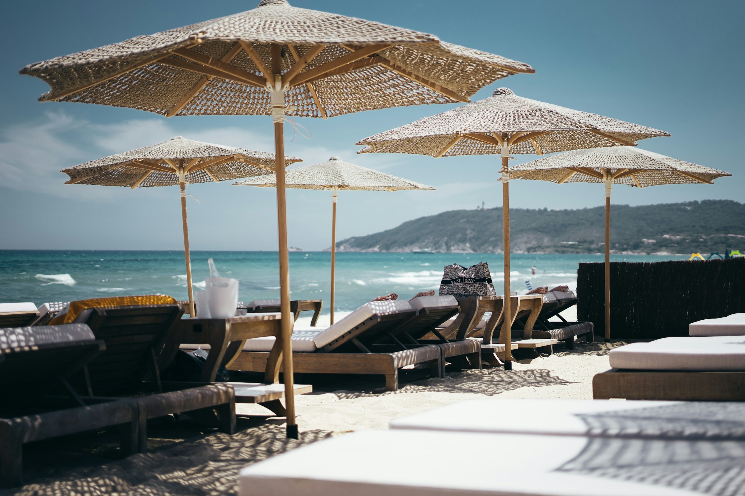 Wicker umbrellas and sun loungers on a sandy beach overlooking the ocean, with hills in the background, under a clear blue sky.