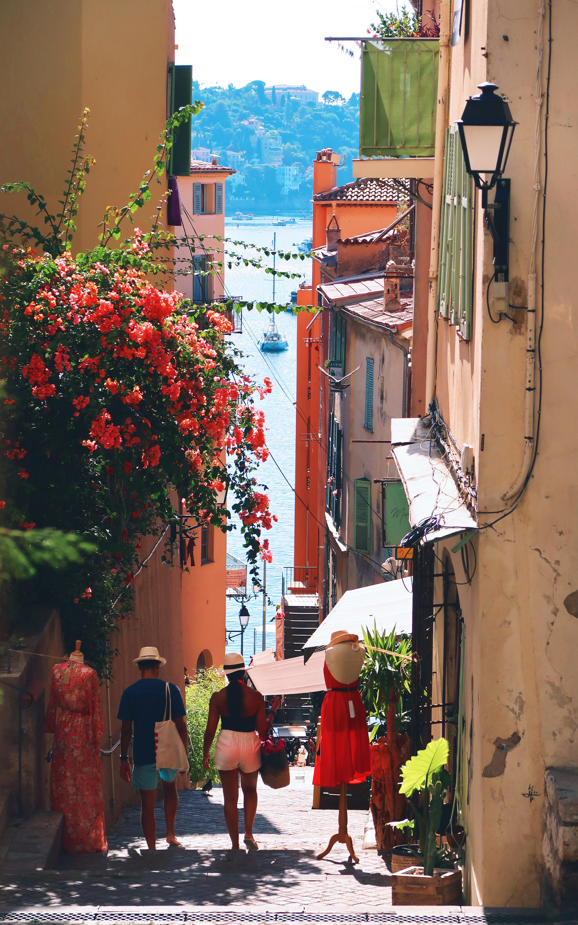 Narrow picturesque alley in a Mediterranean coastal village, with colorful buildings, flowering plants, and people strolling toward the sea.