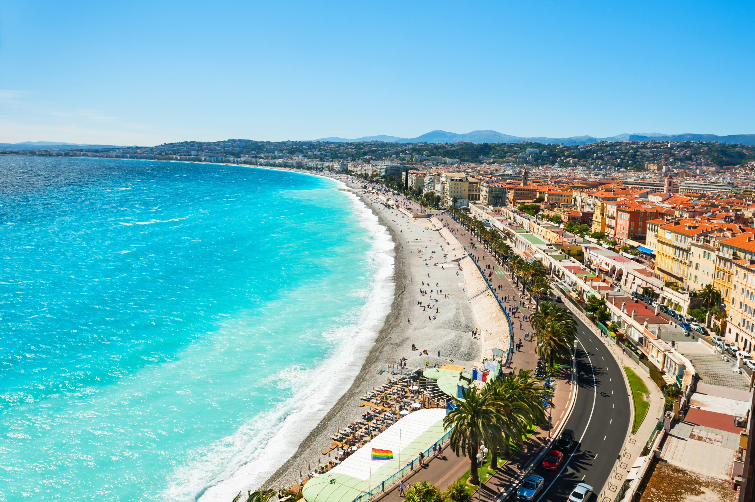 Luftfoto af Nice strandpromenade med turkisblåt hav og solrige badestrande langs kysten, samt byen Nice i baggrunden under en klar blå himmel