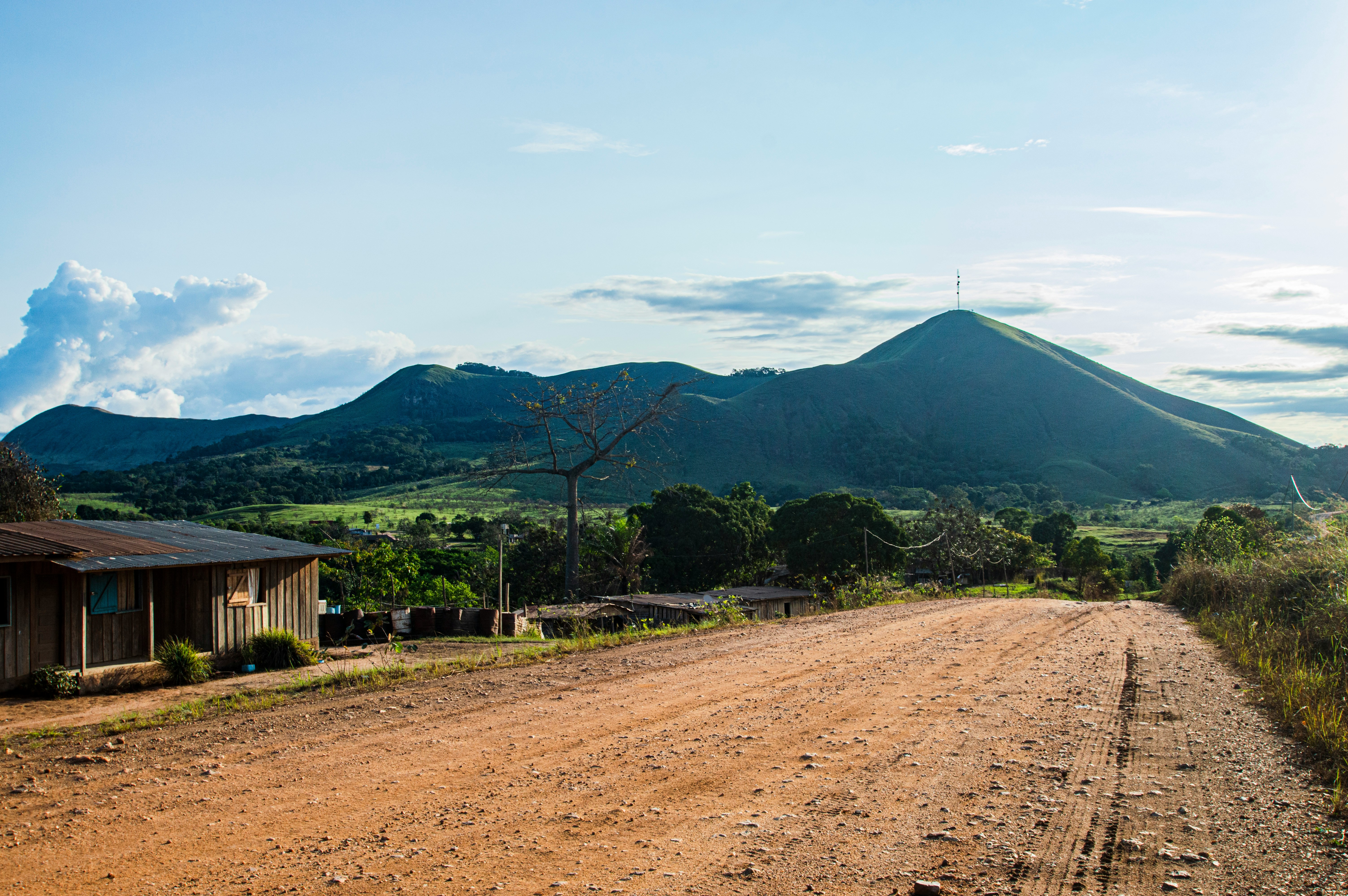 Travel to Gabon - road leading to lush green hills under a clear blue sky with scattered clouds, rustic wooden house on the left.
