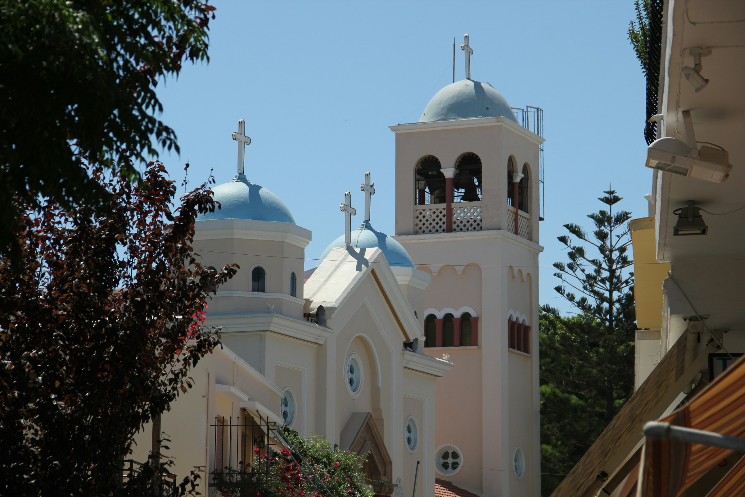White church with blue domes and a bell tower, surrounded by trees and buildings, under a clear blue sky.