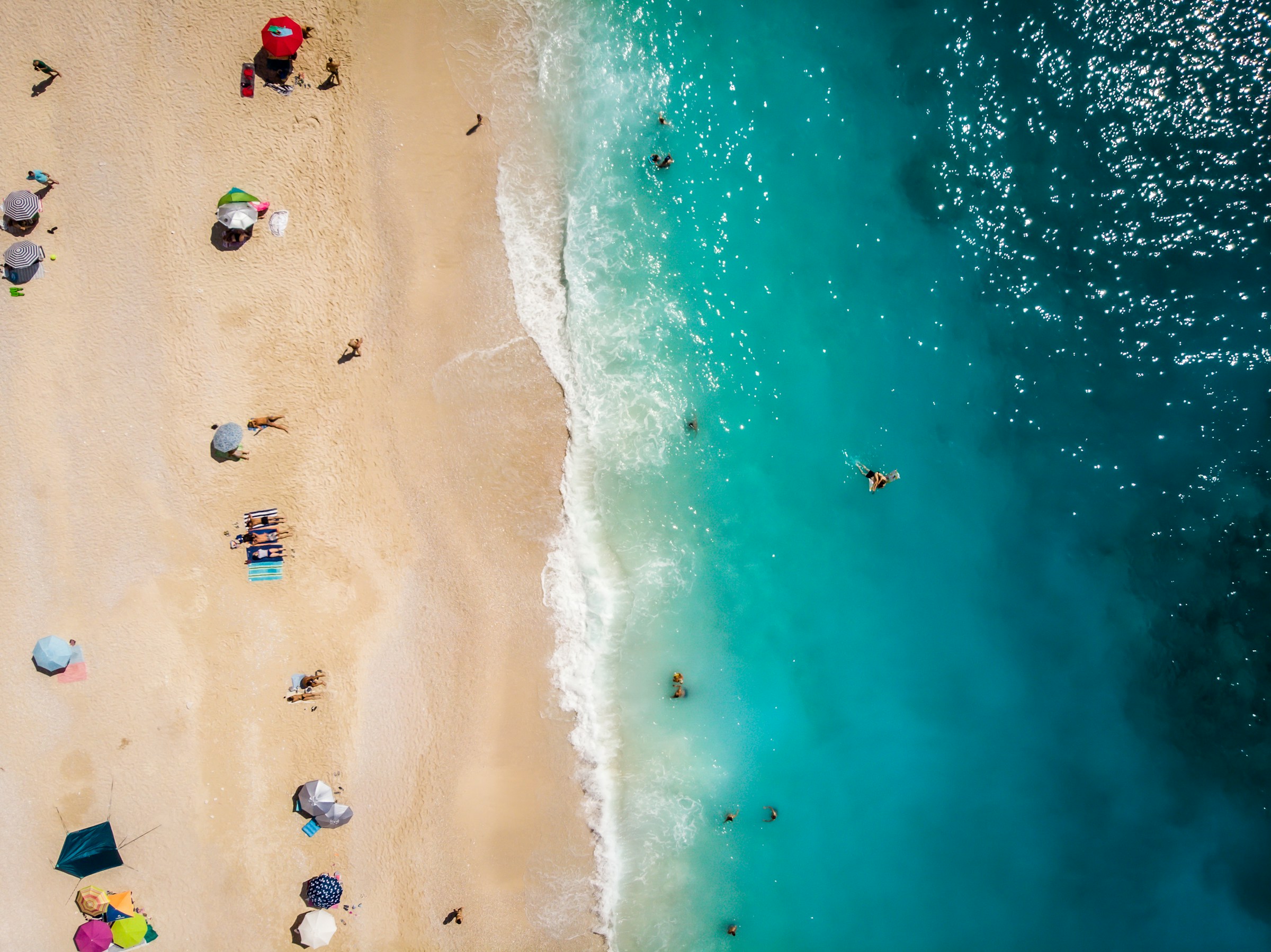 Aerial view of a beach with turquoise ocean water and sandy shore, dotted with sunbathers and colorful umbrellas.
