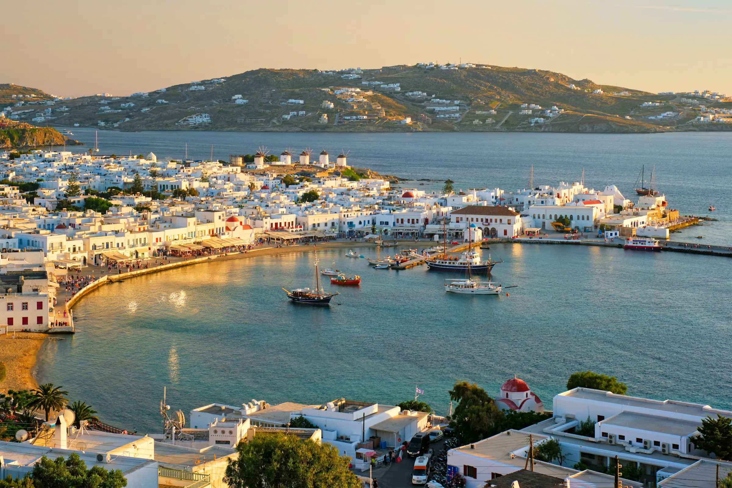 Kystlandskab af Mykonos, Grækenland ved solnedgang, med traditionelle hvide bygninger, både i havnen og bakker i baggrunden