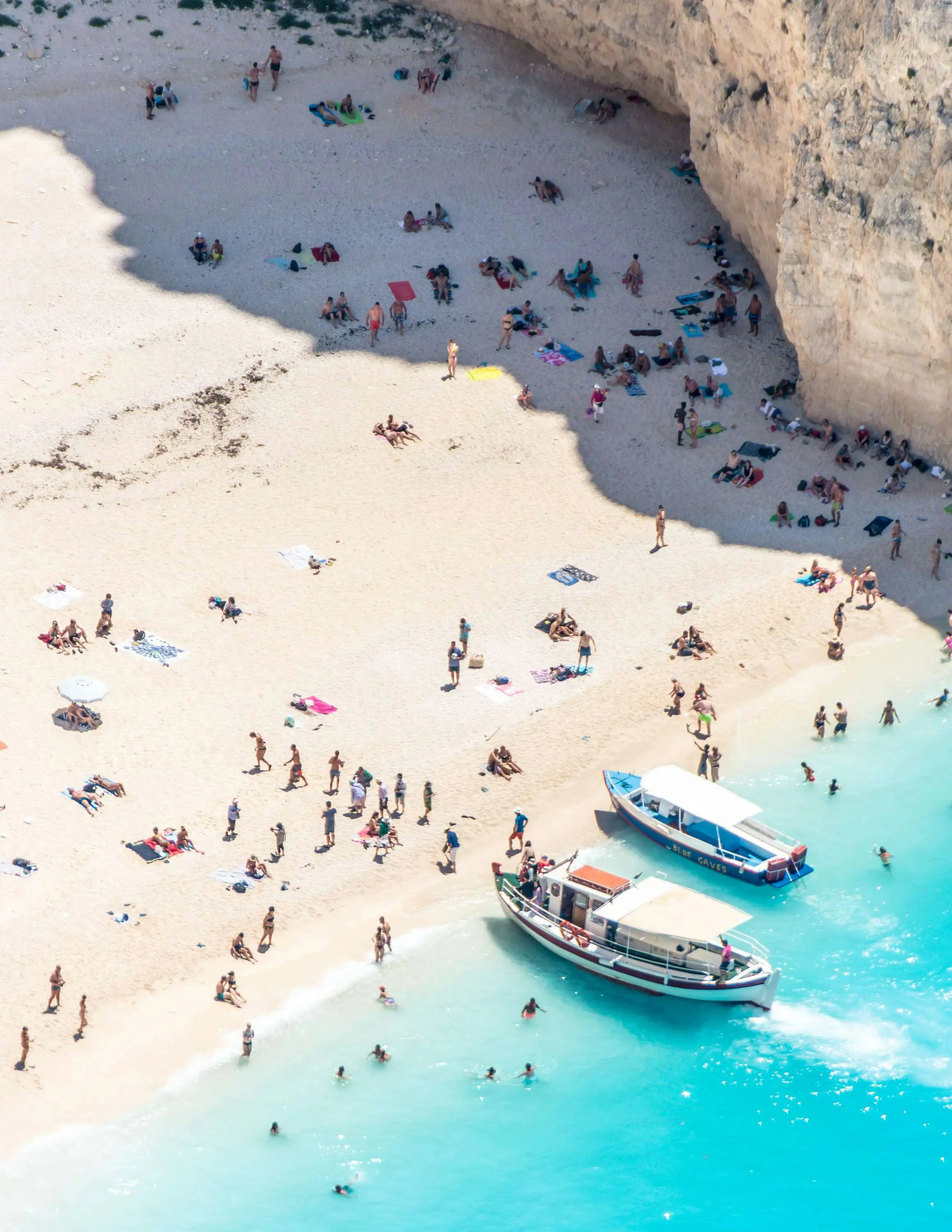 Strand med klipper, fyldt med solbadere, besøgende både og turkisblåt hav