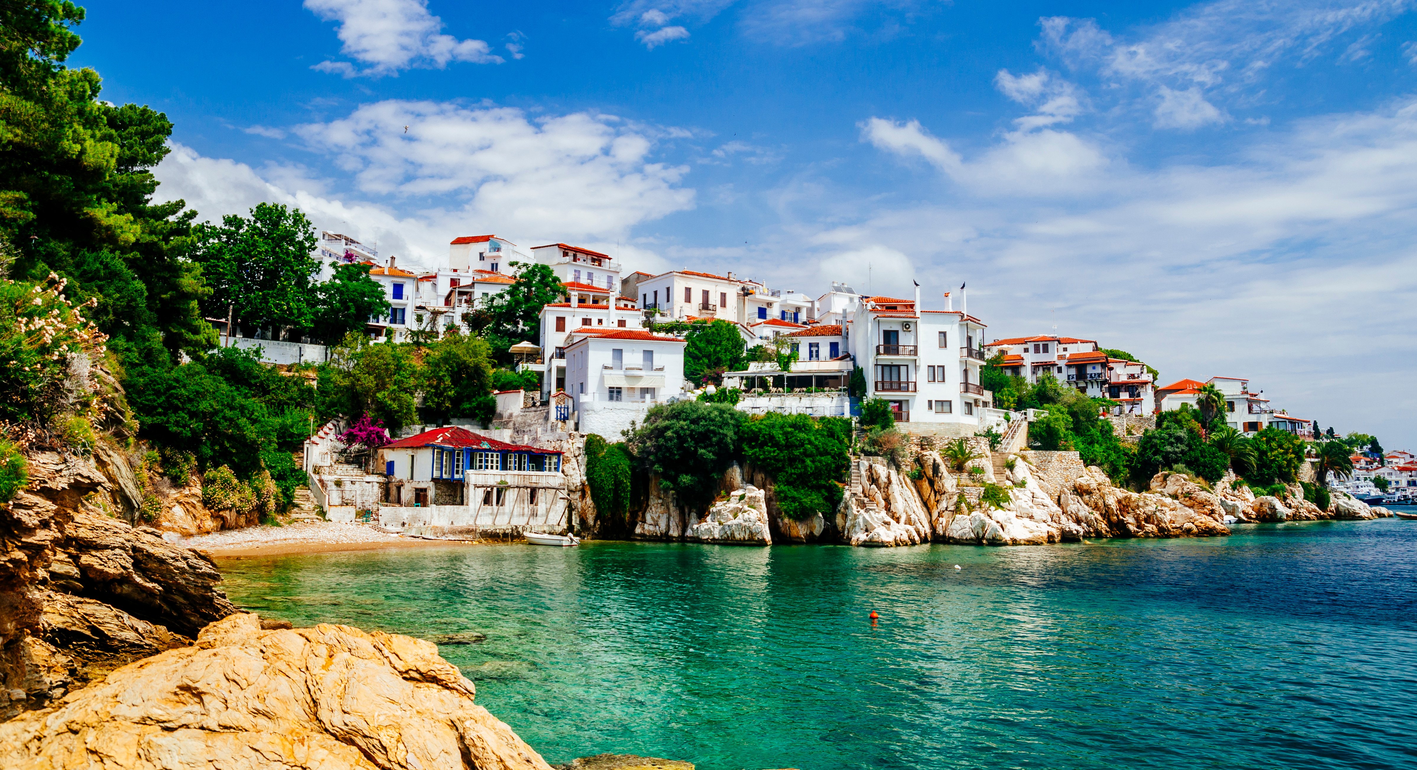Travel to Skiathos - Coastal village with white buildings and red roofs on rocky cliffs overlooking clear turquoise sea under a blue sky.
