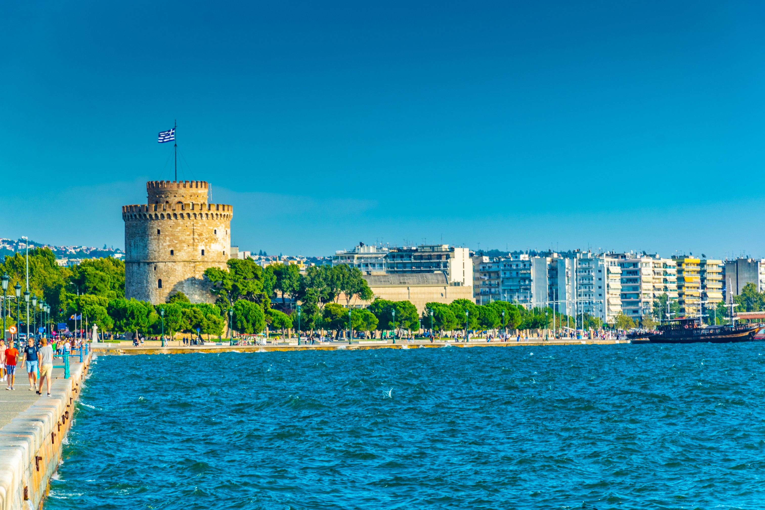 Hvidtårn i Thessaloniki med græsk flag, omgivet af hav og byens skyline i solskinsvejr