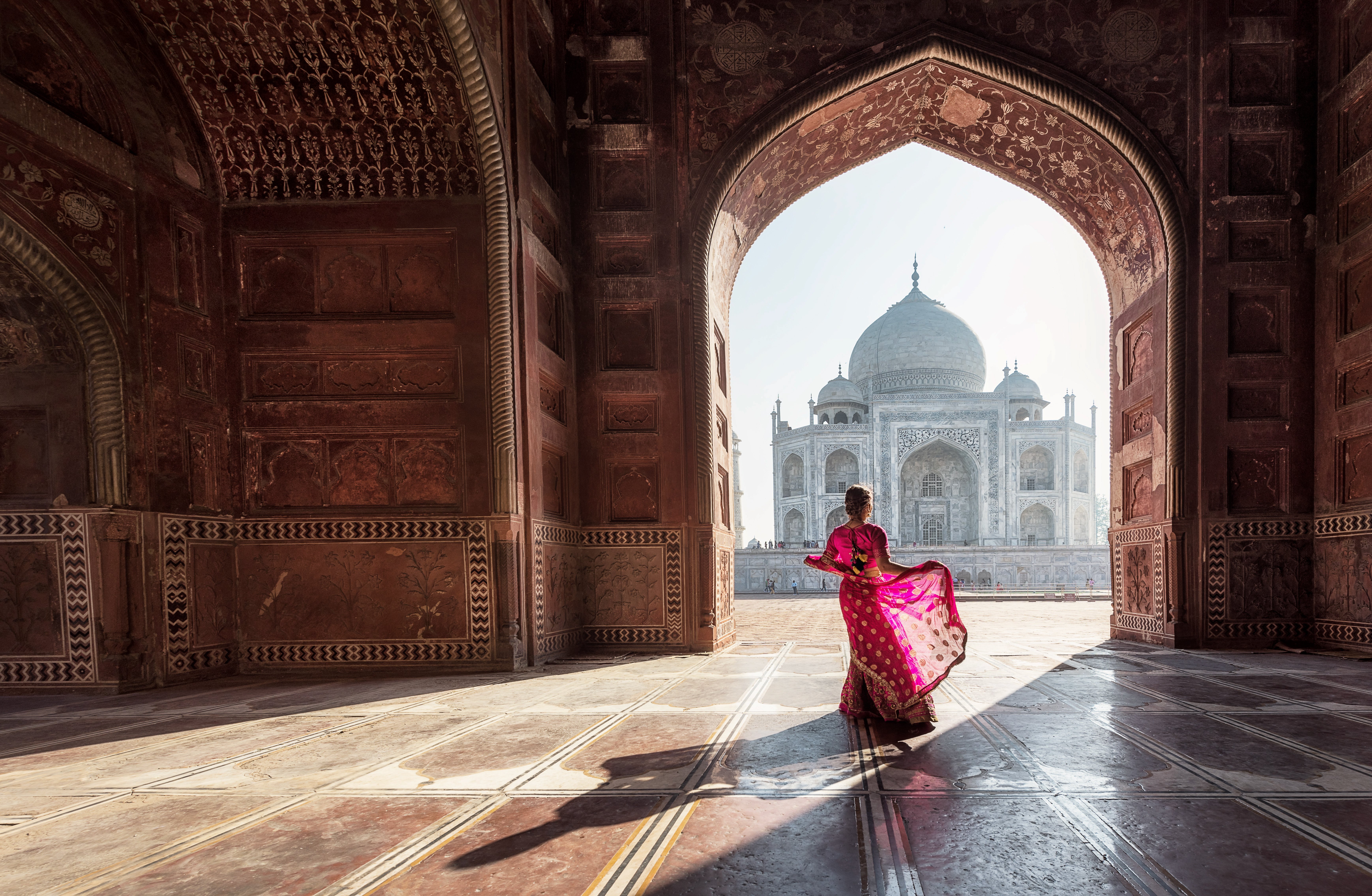 Kvinde i farverig sari står i en buegang og kigger mod Taj Mahal, Agra, Indien, under klart sollys