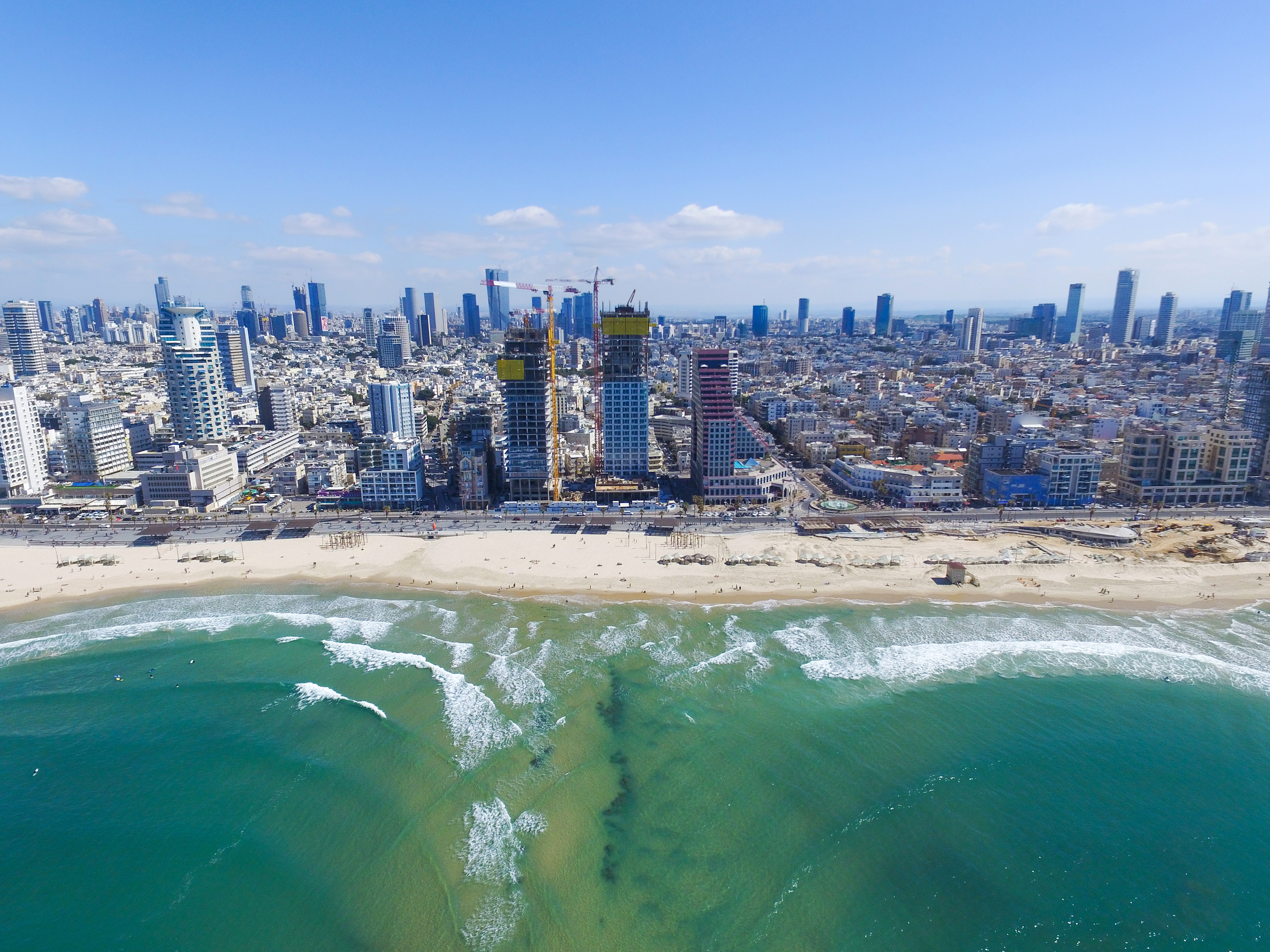 Luftfoto af bylandskab ved kysten med højhuse, strand og bølger i forgrunden under en klar blå himmel i Tel Aviv