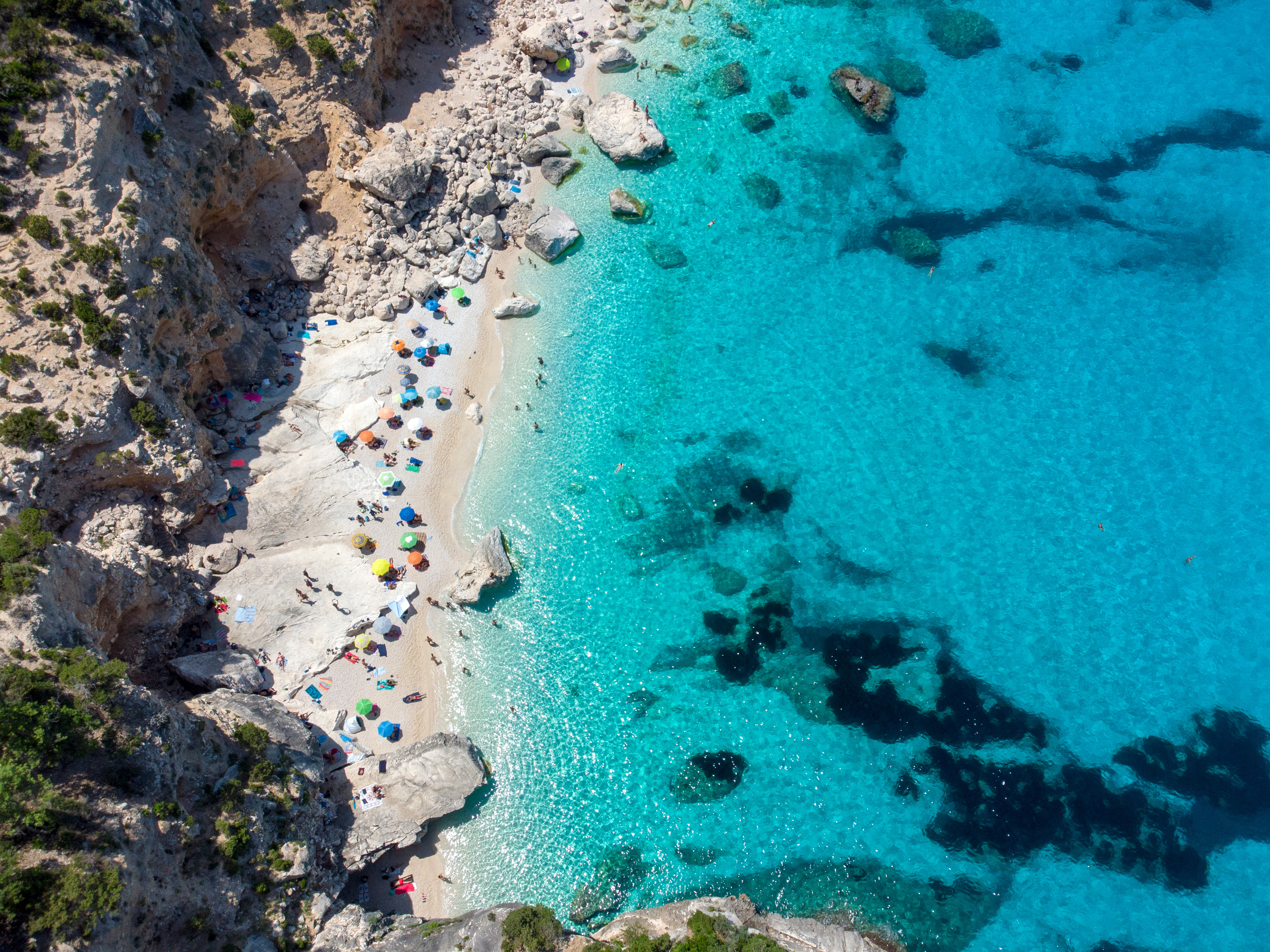 Luftfoto af en smuk turkisblå strand med klipper, hvor folk ligger under farverige parasoller i solen
