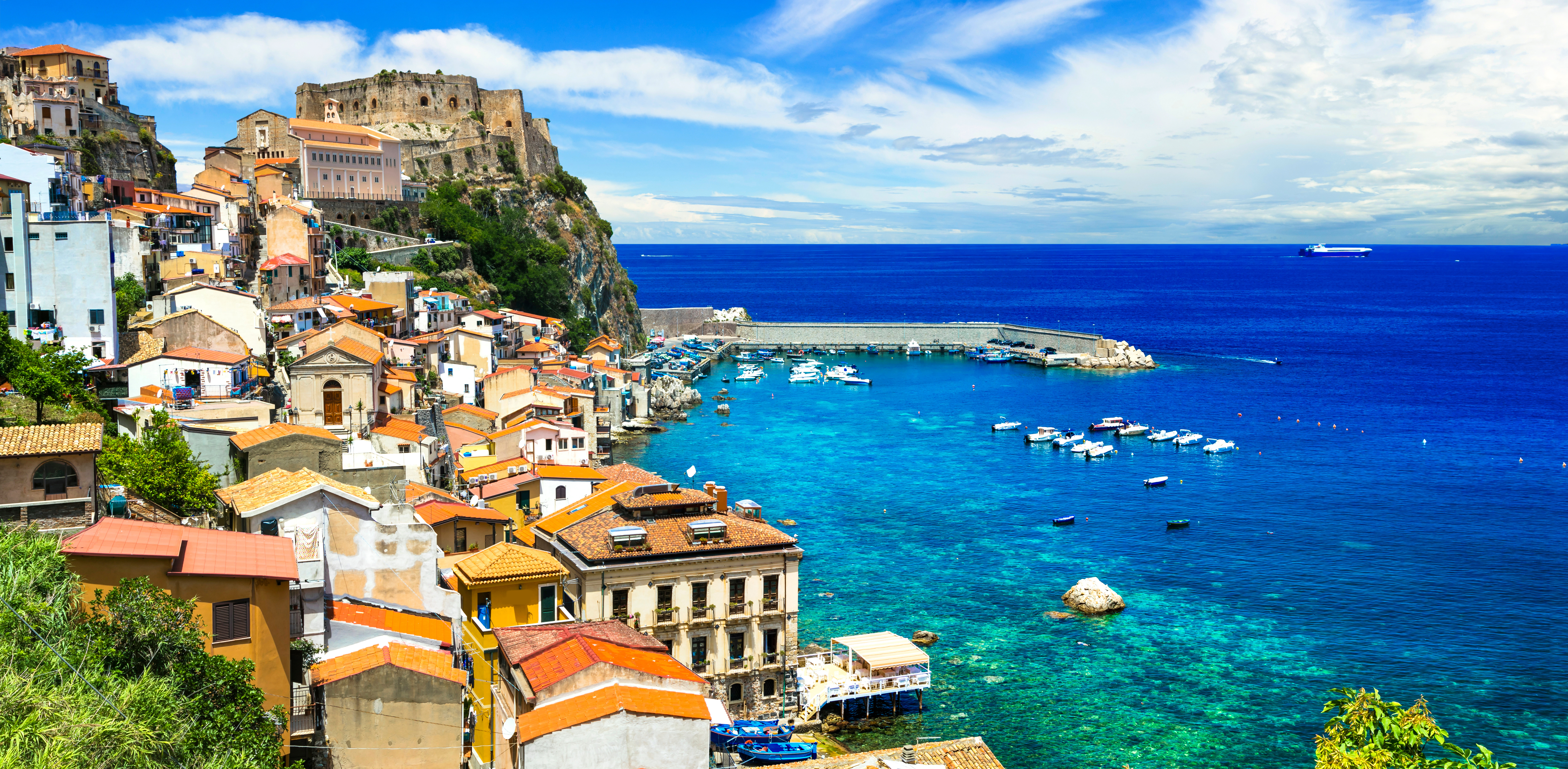 Kystlandskab i Italien med farverige huse ved havet, en smuk fæstning på en klippetop og både i det klare blå vand under en klar himmel