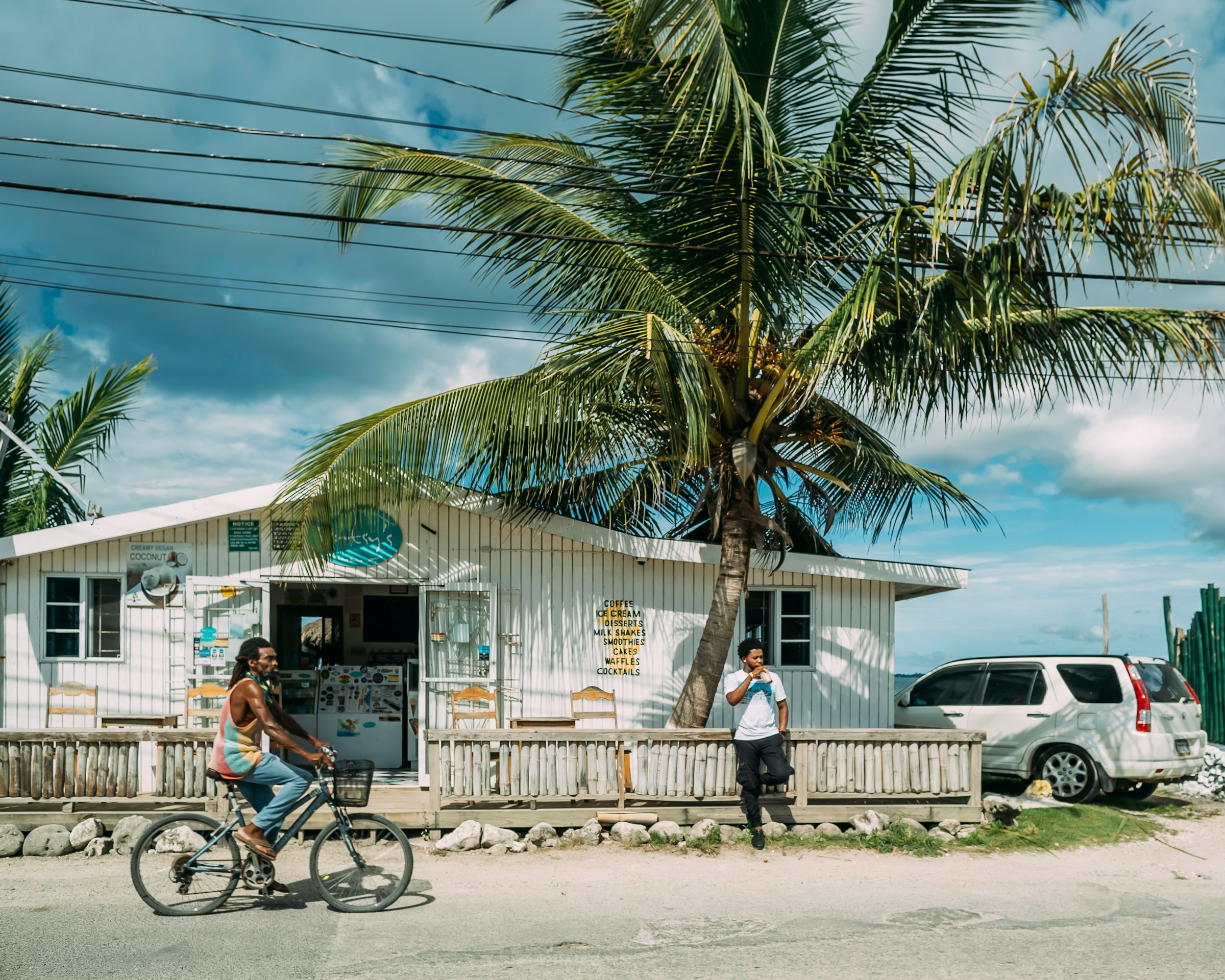 Tropical café with palm trees, a person riding a bicycle, and another person standing outside; a parked SUV is visible nearby.