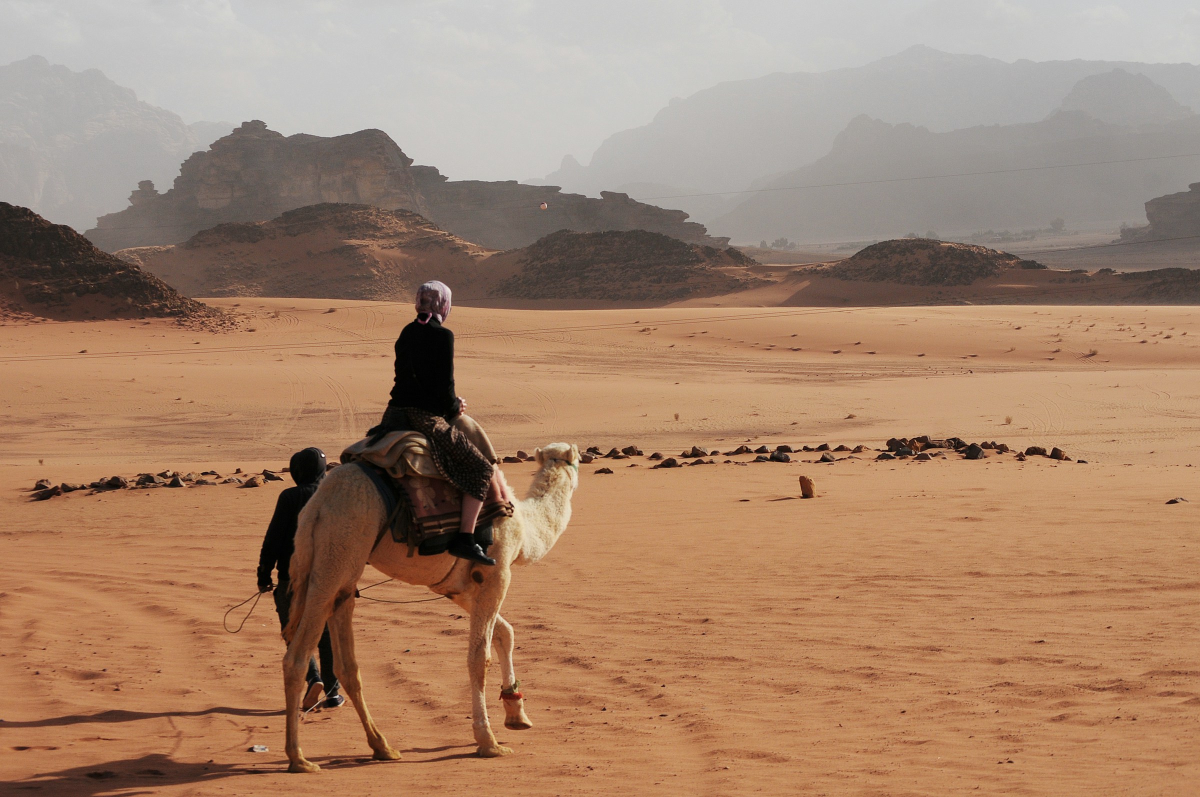 Person riding a camel in the desert with rocky hills in the background, under a clear sky.