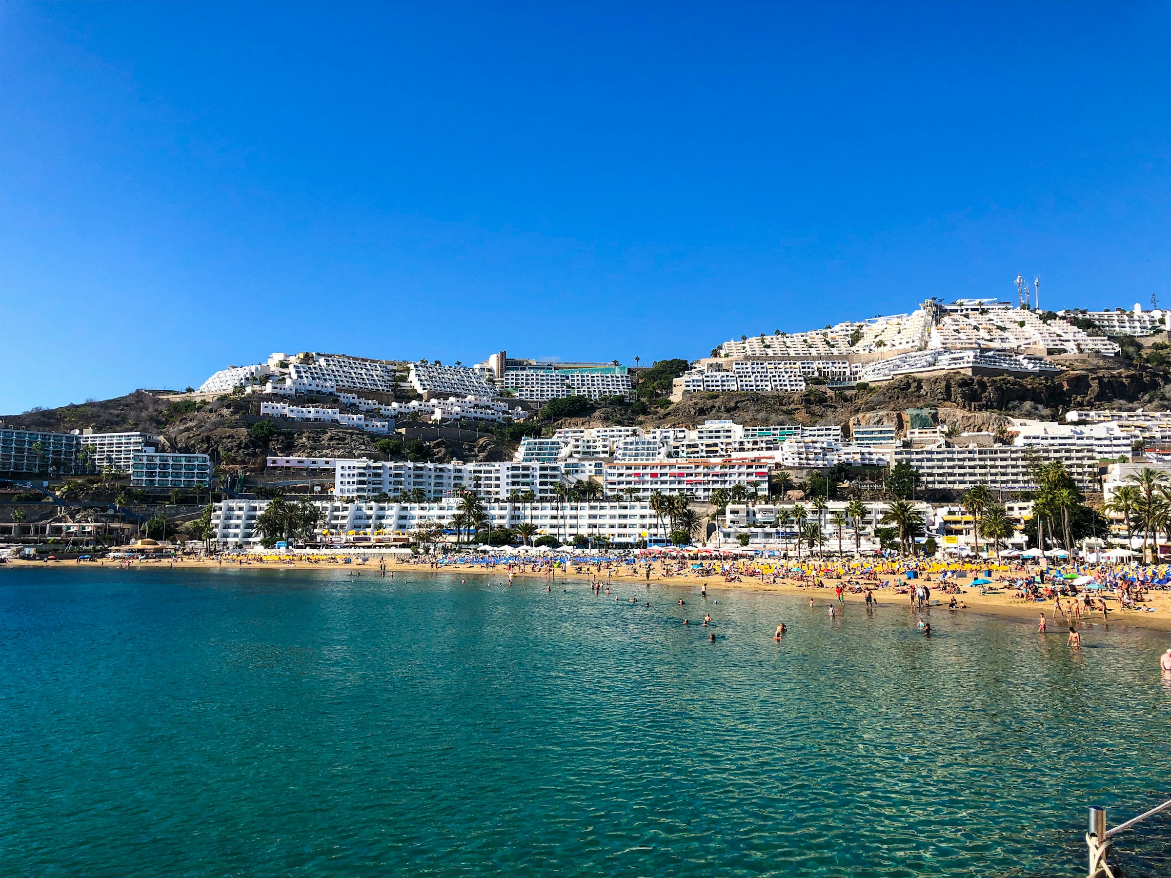 Strand og resort på Gran Canarias kyst, De Kanariske Øer, Spanien, med turkisblåt hav, sandstrand og hvide højhuse på en bakke under en klar blå himmel.