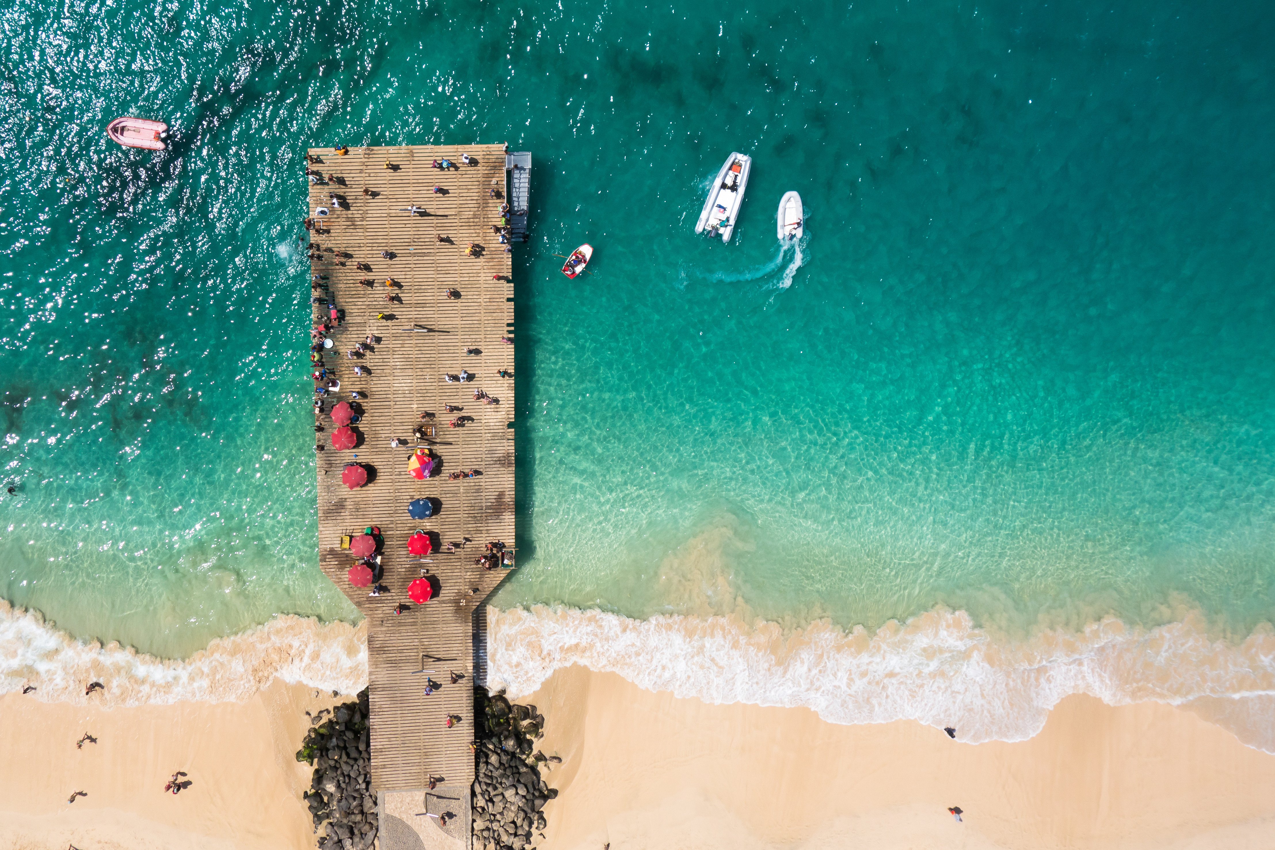 Luftfoto af badebro ved strand med turkisblåt vand og både, besøgt af mennesker under farverige parasoller