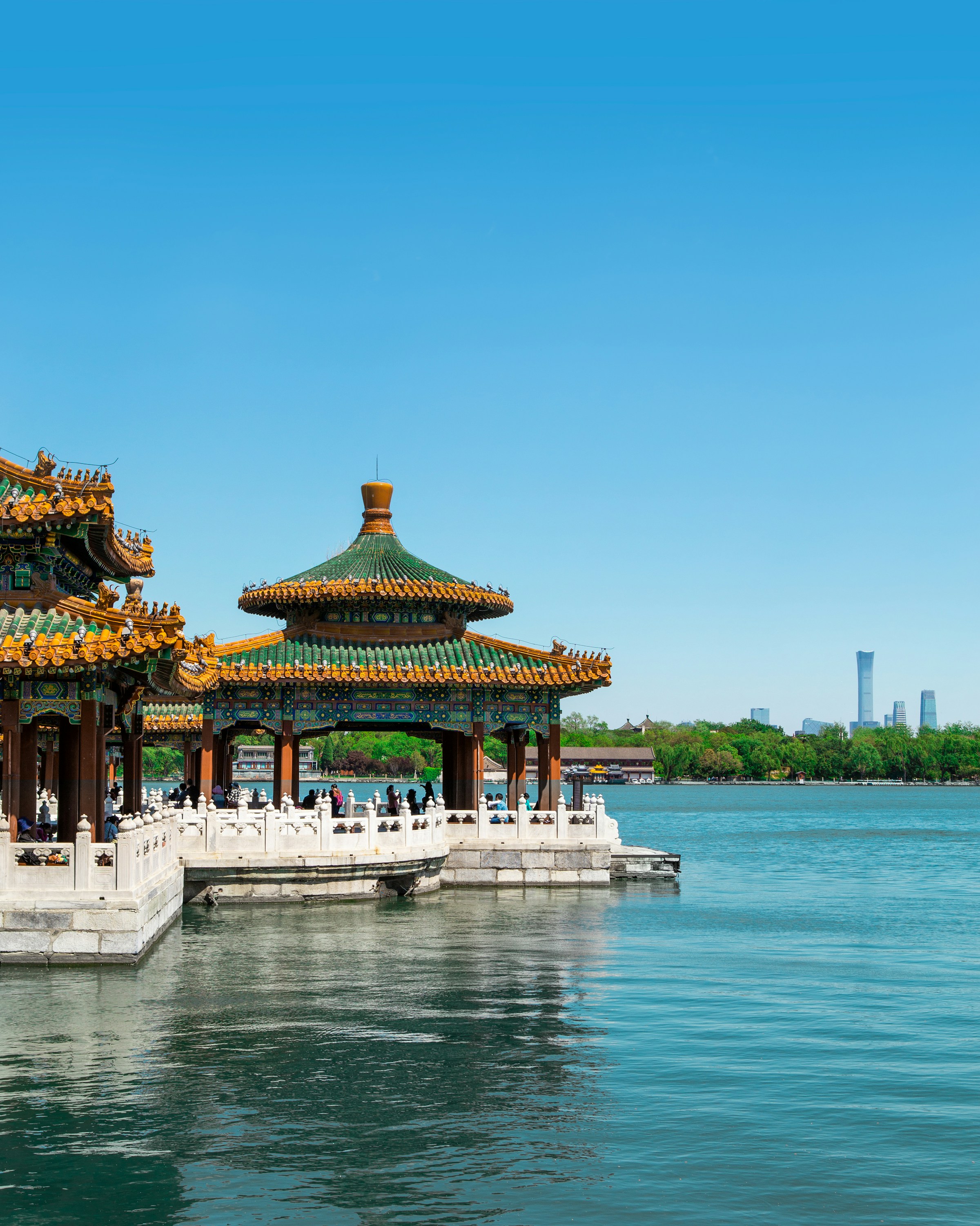 Traditional Chinese pavilion overlooking a serene lake under a clear blue sky, with a city skyline visible in the distance